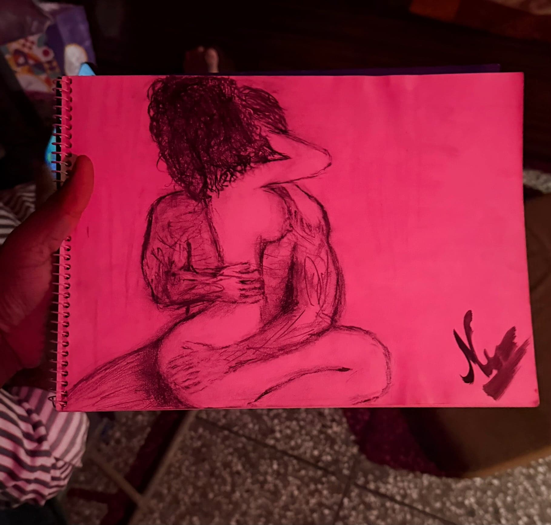 A charcoal drawing on bright pink paper of two figures in a close embrace seen from behind, one with dark curly hair, signed with a bold N at the lower right, in a spiral-bound sketchbook held up by a hand in a dimly lit room