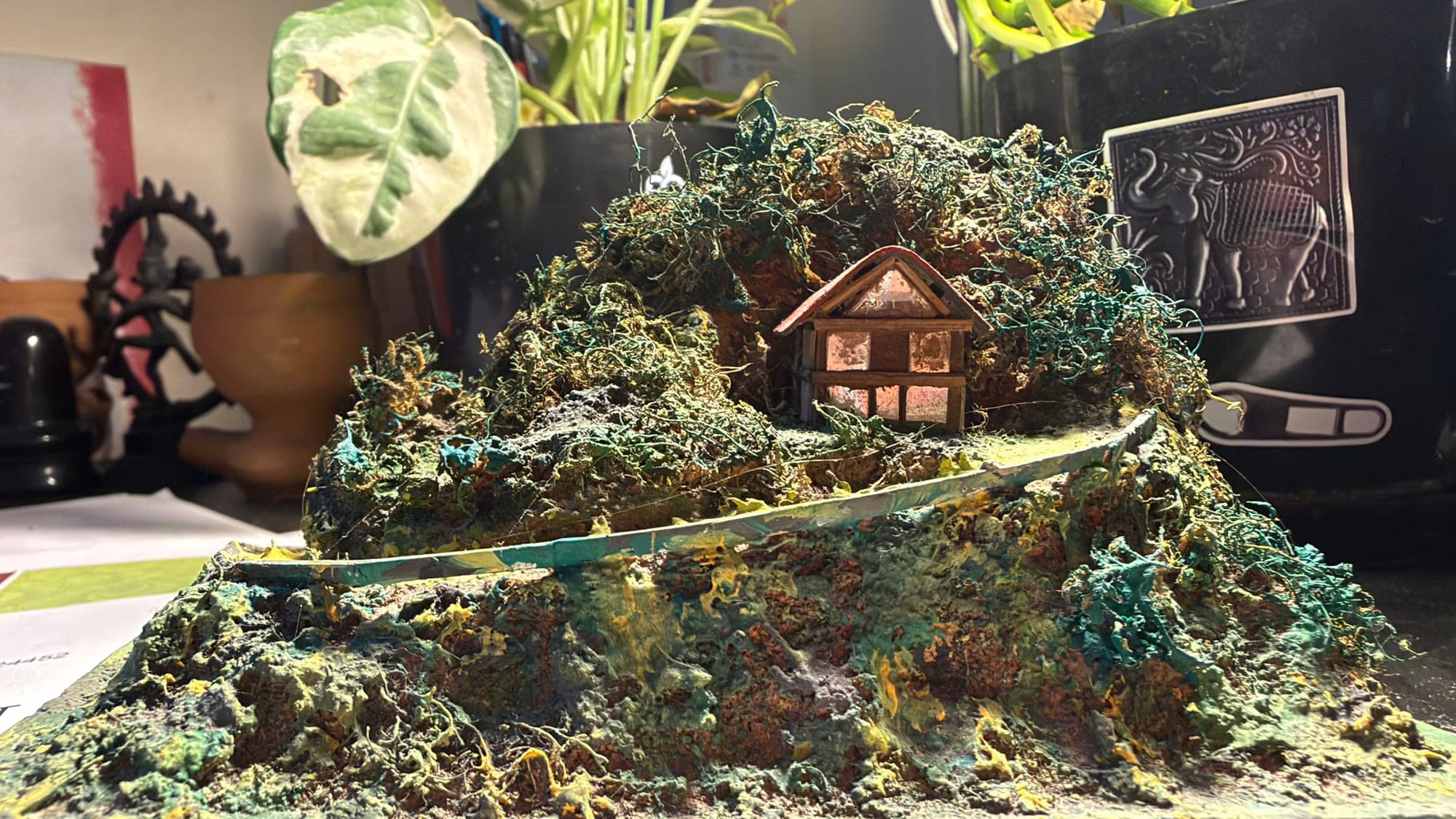 A hand-made miniature hill in greens, teal and rust, with a small timber house with a red roof and softly glowing windows, and a thin curved road winding round it; plants and a black planter stand behind it on a desk