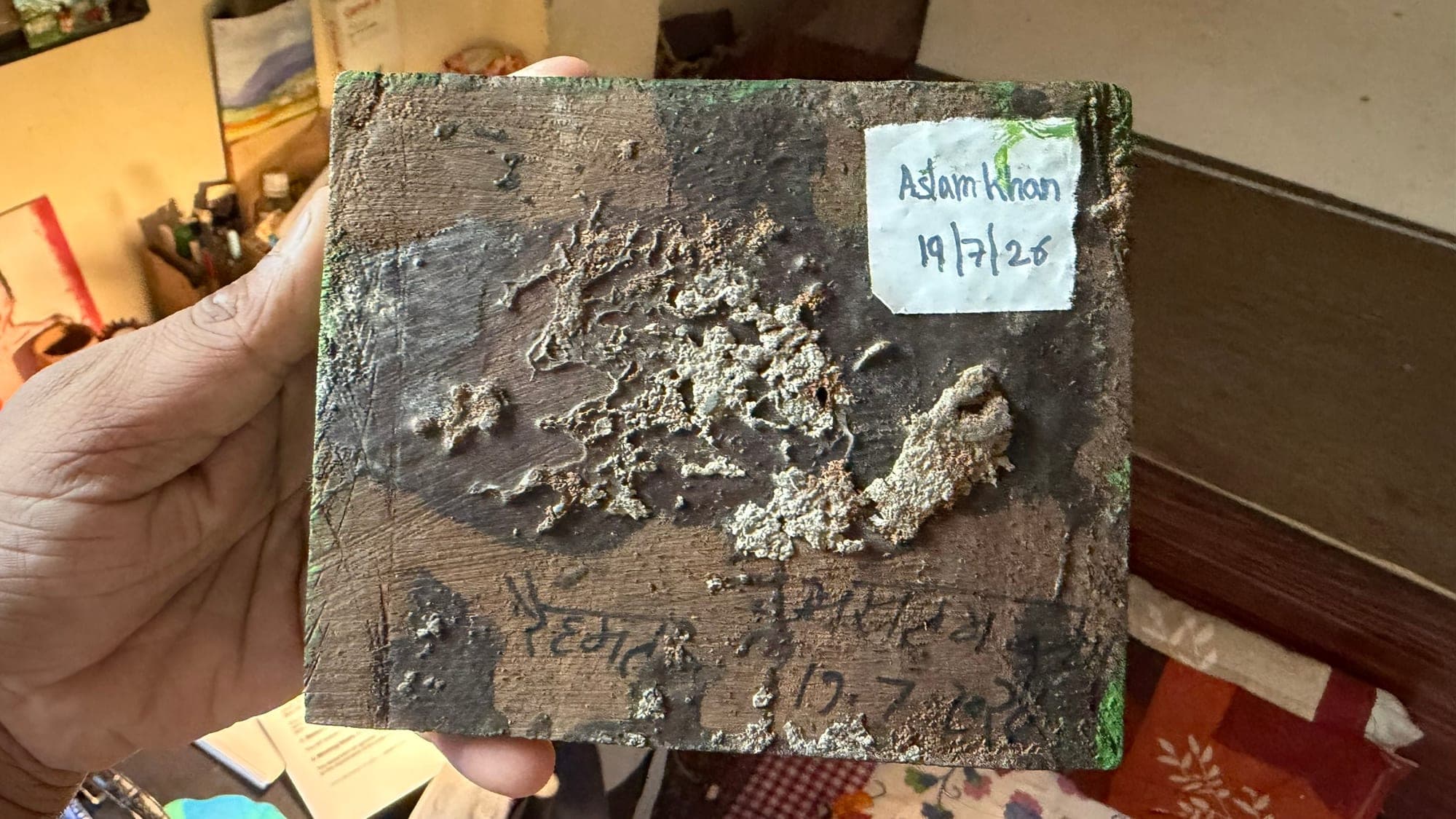 A hand holding the back of a small wooden panel, dark and textured with pale crusted growth, with a white paper label reading Aslam Khan 19/7/26 in one corner and handwriting along the bottom edge