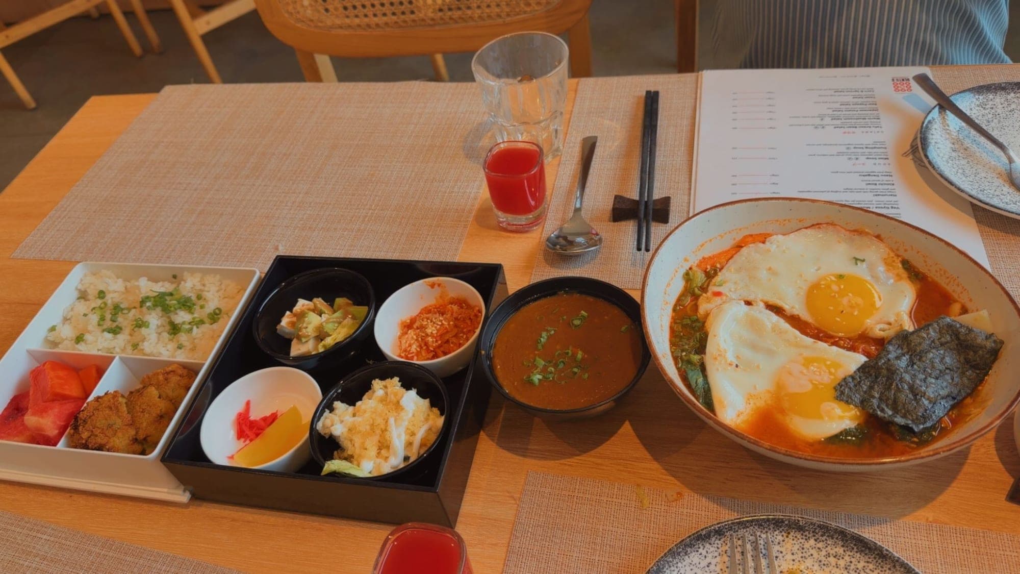 A table seen from above: a black bento box of small bowls of pickles, salad, avocado and kimchi beside a white tray of rice, fritters and watermelon, a bowl of dark curry, two red drinks, chopsticks on a rest, and a bowl of ramen with two fried eggs at the right