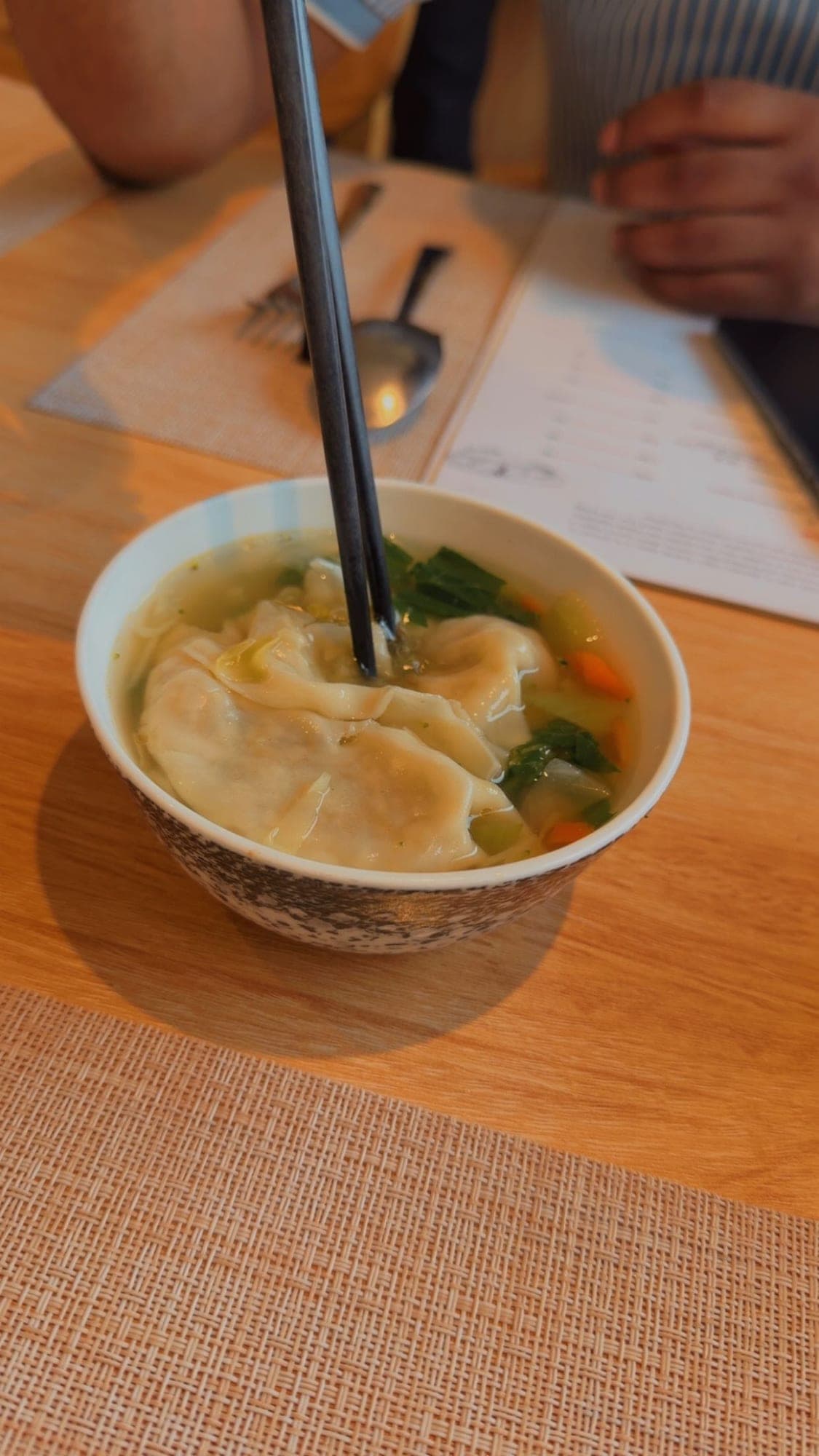A white bowl of clear broth with large dumplings, bok choy and carrot, black chopsticks dipping into the soup, on a wooden table with a woven placemat and a menu