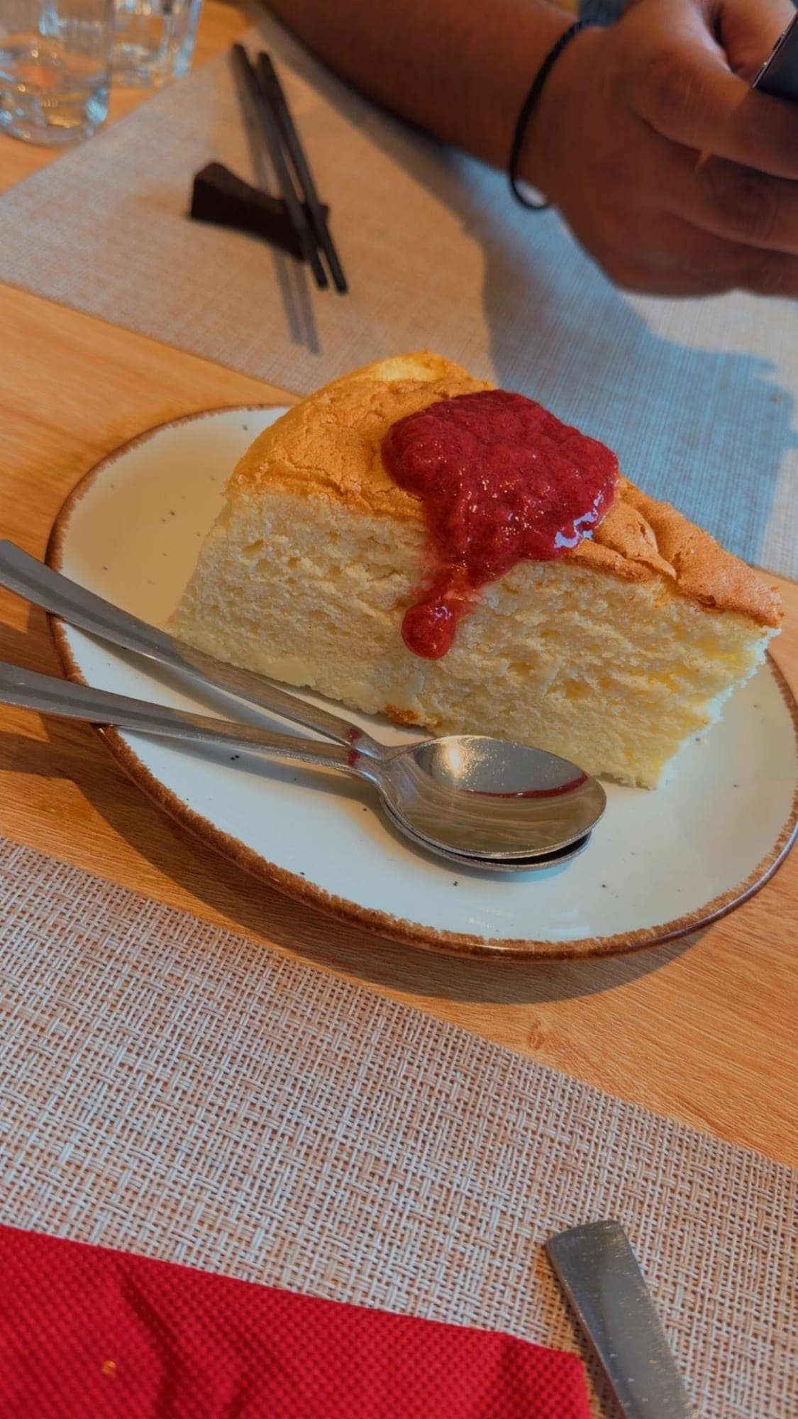 A thick wedge of golden, airy Japanese cheesecake with a spoonful of red berry sauce on top, on a speckled plate with two spoons, on a wooden table with woven placemats and a red napkin
