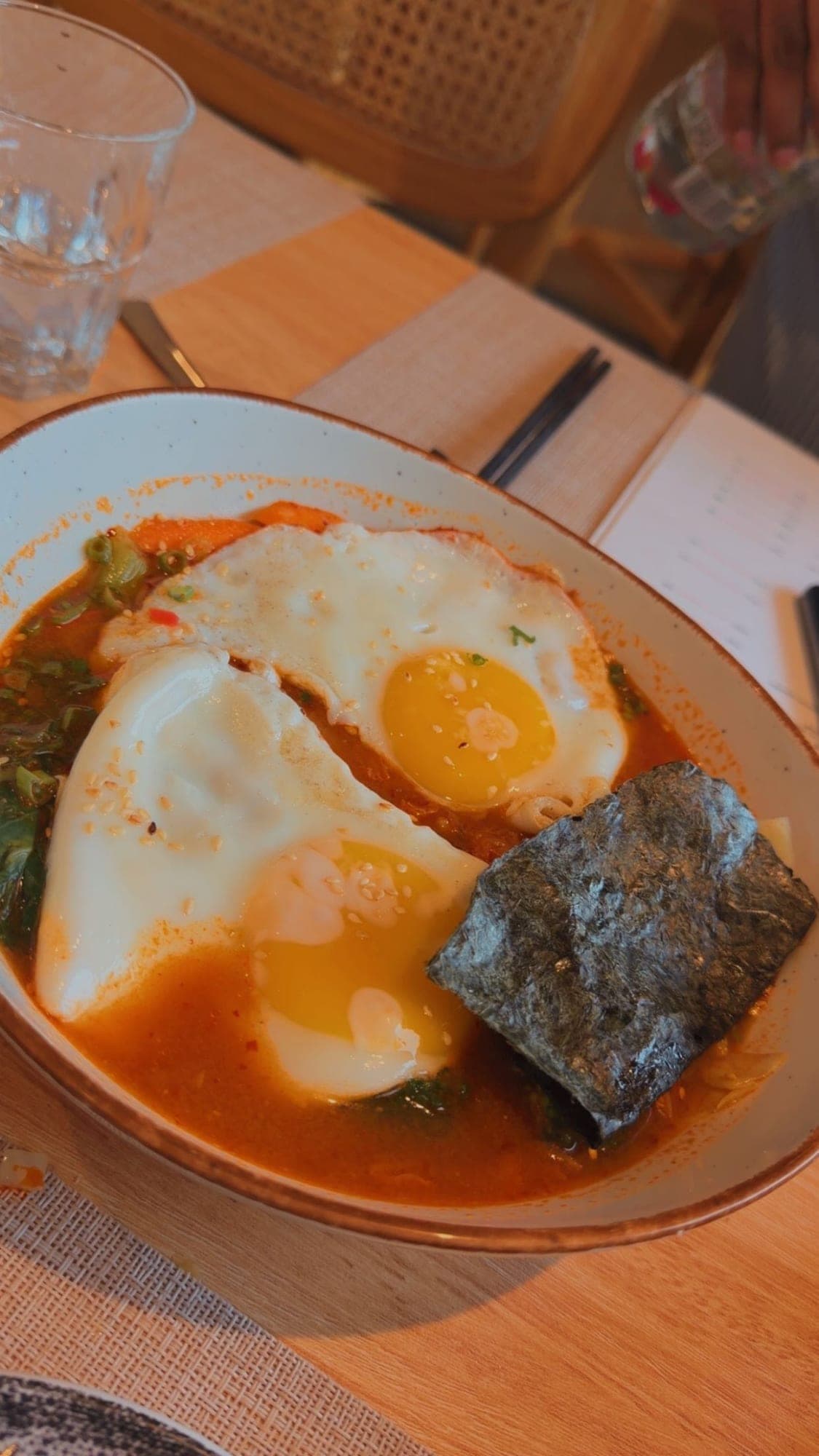 A wide speckled bowl of orange-red broth topped with two fried eggs with sesame seeds, spring onion and a sheet of dark nori, on a wooden table with woven placemats, a water glass and chopsticks beyond