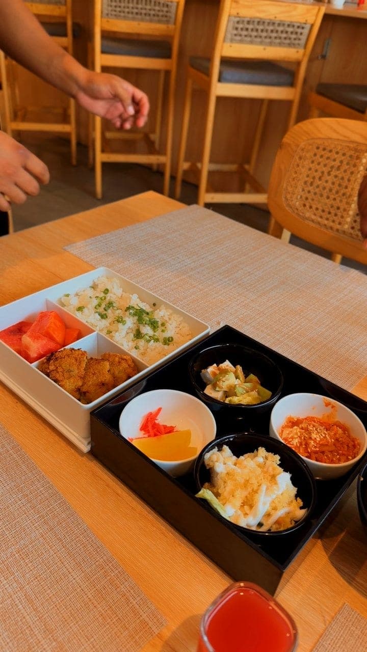 An opened bento box on a wooden table: a white tray of rice, fritters and watermelon beside a black box of small bowls, a hand lifting the black lid away at the top, and a red drink at the lower right