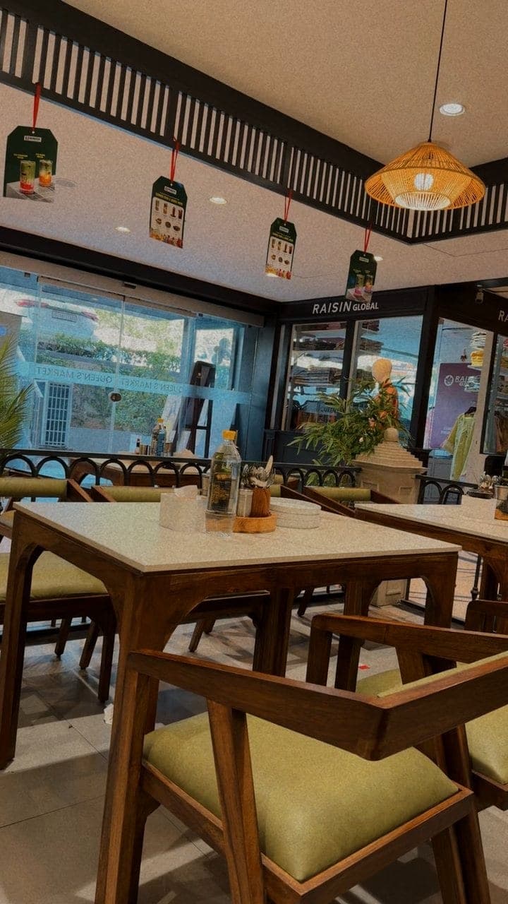 The inside of a bright restaurant: wooden chairs with green seats at white-topped tables in the foreground, a woven hanging lamp and small tags hanging from a dark slatted ceiling, and tall windows with plants at the back