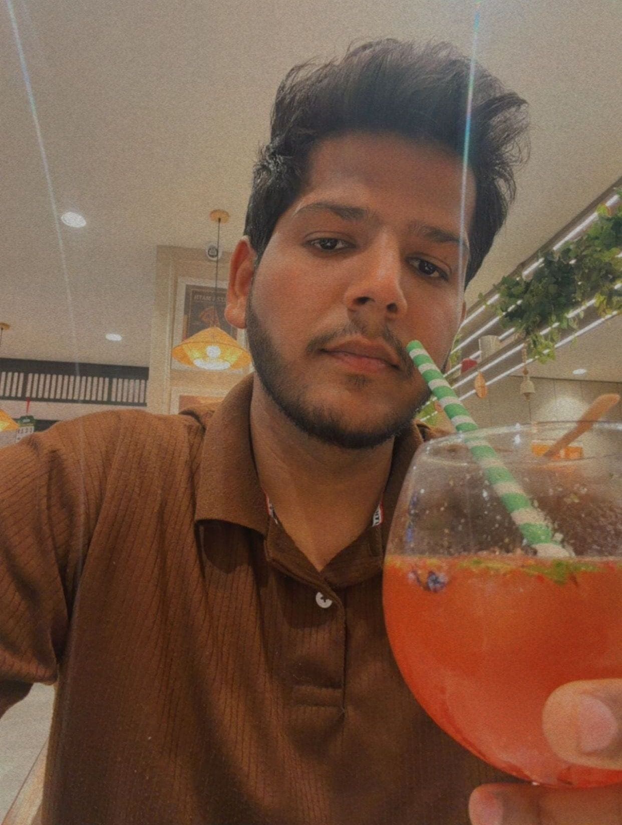 A selfie of a bearded man in a brown ribbed polo shirt holding a large round glass of orange-red mojito with a green striped straw up to the camera, in a warm restaurant with a hanging woven lamp and plants along the ceiling
