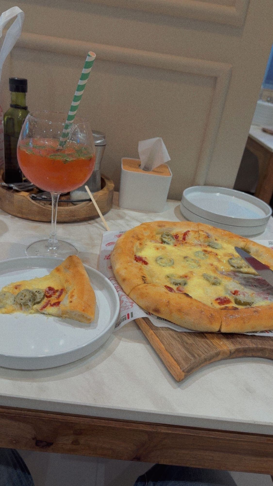 A round cheese pizza with strips of red pepper and sliced green jalapeños on a wooden paddle over printed paper, one slice cut onto a white plate at the left, beside a tall stemmed glass of orange-red mojito with a green striped paper straw, on a white marble table