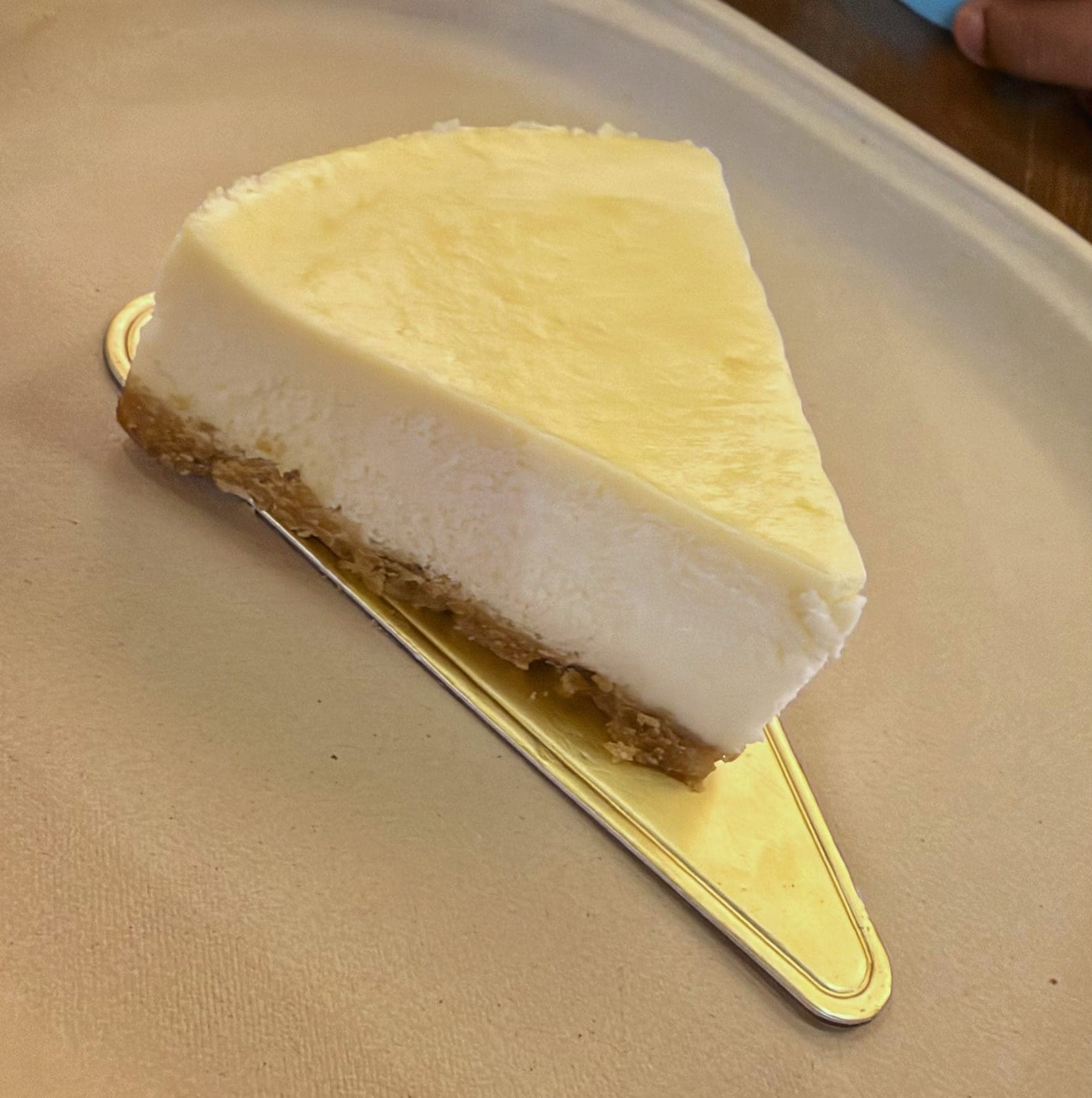 A triangular slice of plain baked cheesecake with a pale golden top and a biscuit crumb base, on a gold cake board on a beige tray
