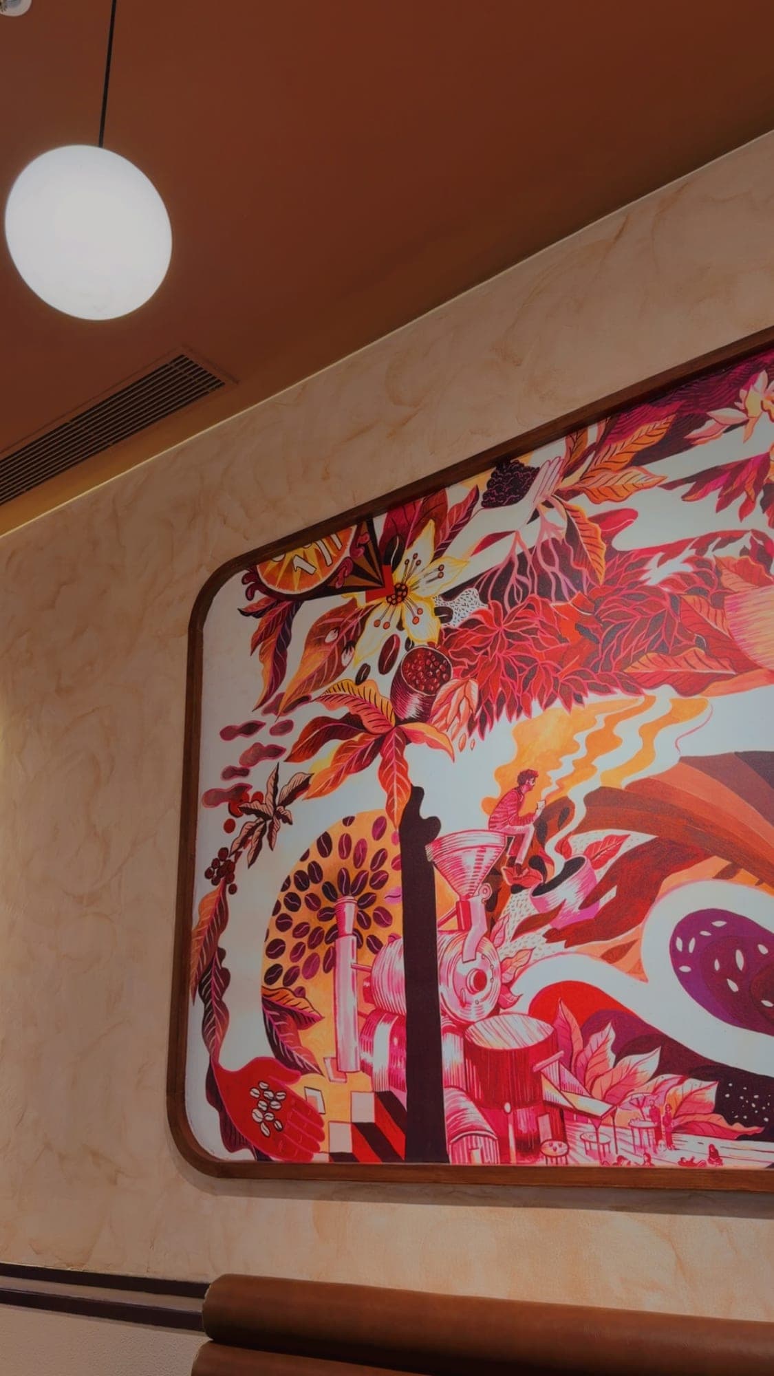 A close view of a framed wall mural in red, pink and orange showing a coffee plant with a white blossom and berries, a large circle of coffee beans, a roasting machine with a small seated figure holding a cup, and curling steam, beside a round white lamp under a terracotta ceiling