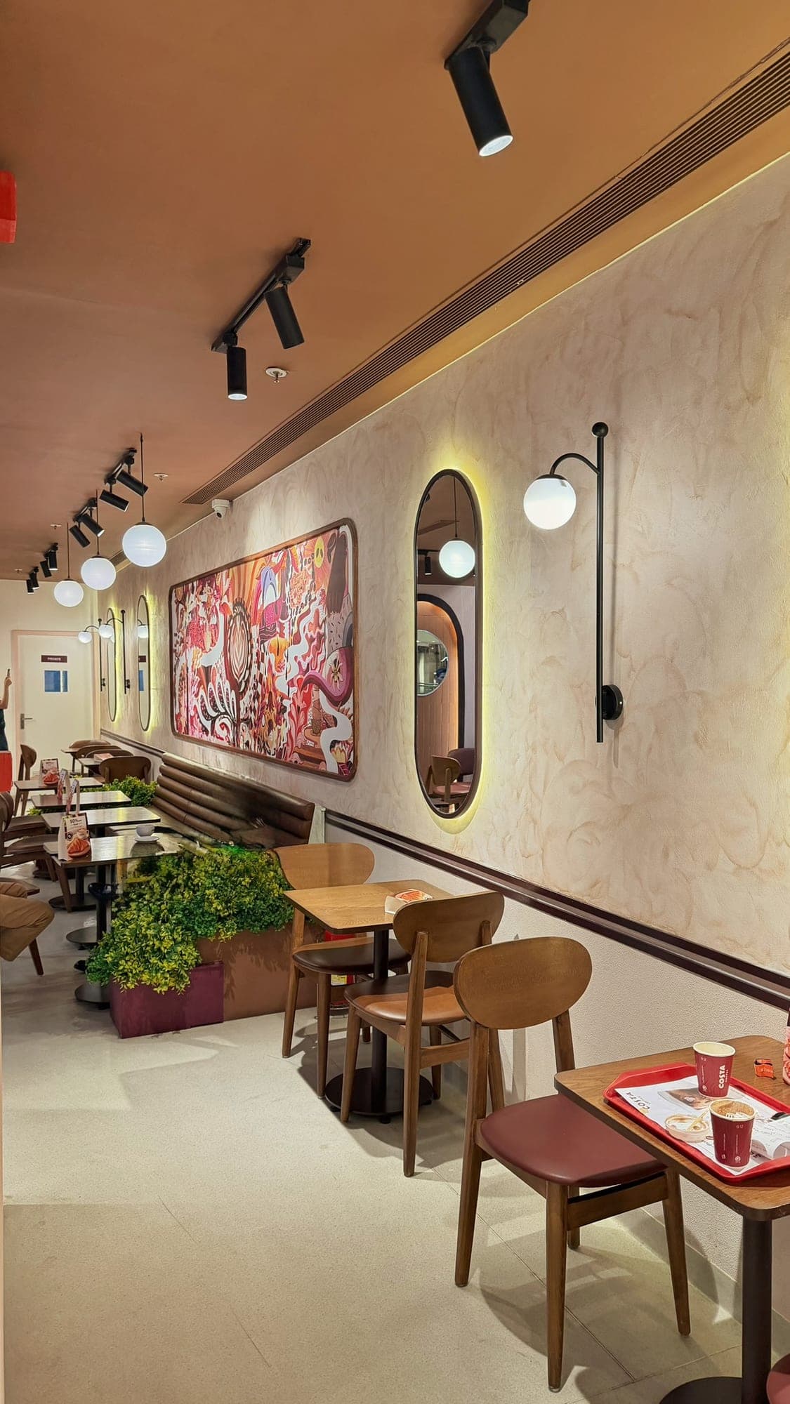 A long café wall of cream plaster under a terracotta ceiling with black track lights and round white globe lamps, holding a large framed mural in red and orange, two tall oval mirrors edged with yellow light, wooden chairs with plum seats, a brown leather bench and a planter of green leaves, with a tray of red Costa cups on a table at the right