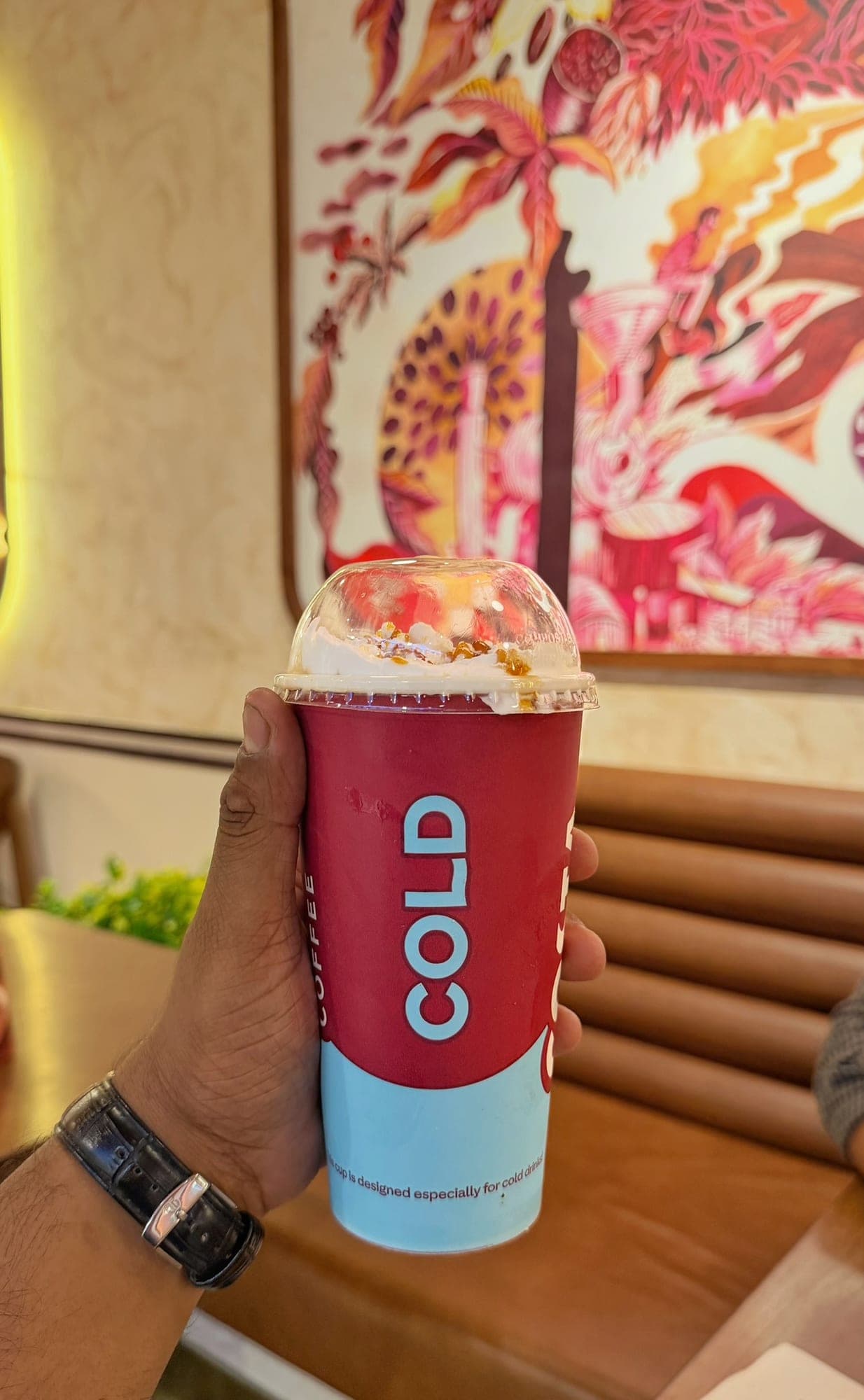 A hand holding a tall red and pale blue paper cup marked COLD COFFEE, topped with whipped cream and caramel under a clear dome lid, in front of a colourful coffee mural and a brown leather booth