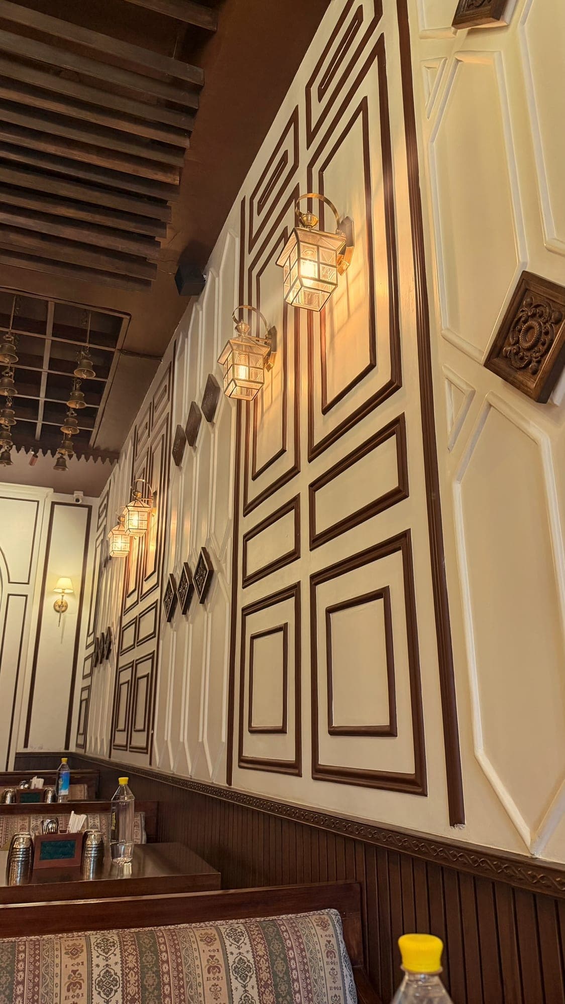 A tall cream wall with dark wood moulded panels, a row of glowing brass lanterns, carved wooden squares set on the diagonal, and a wooden slatted ceiling above, with booths and tables at the bottom