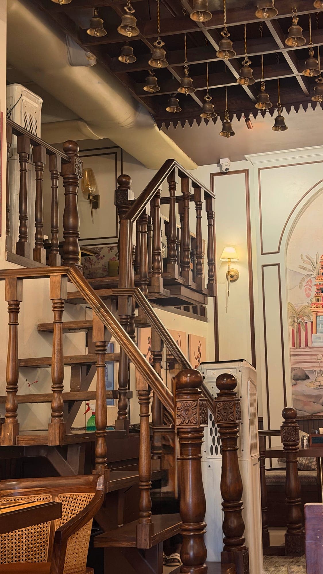 A carved wooden staircase with turned balusters and carved posts climbing to a small upper level under a ceiling of hanging brass bells, with part of the temple mural and a wall lamp at the right