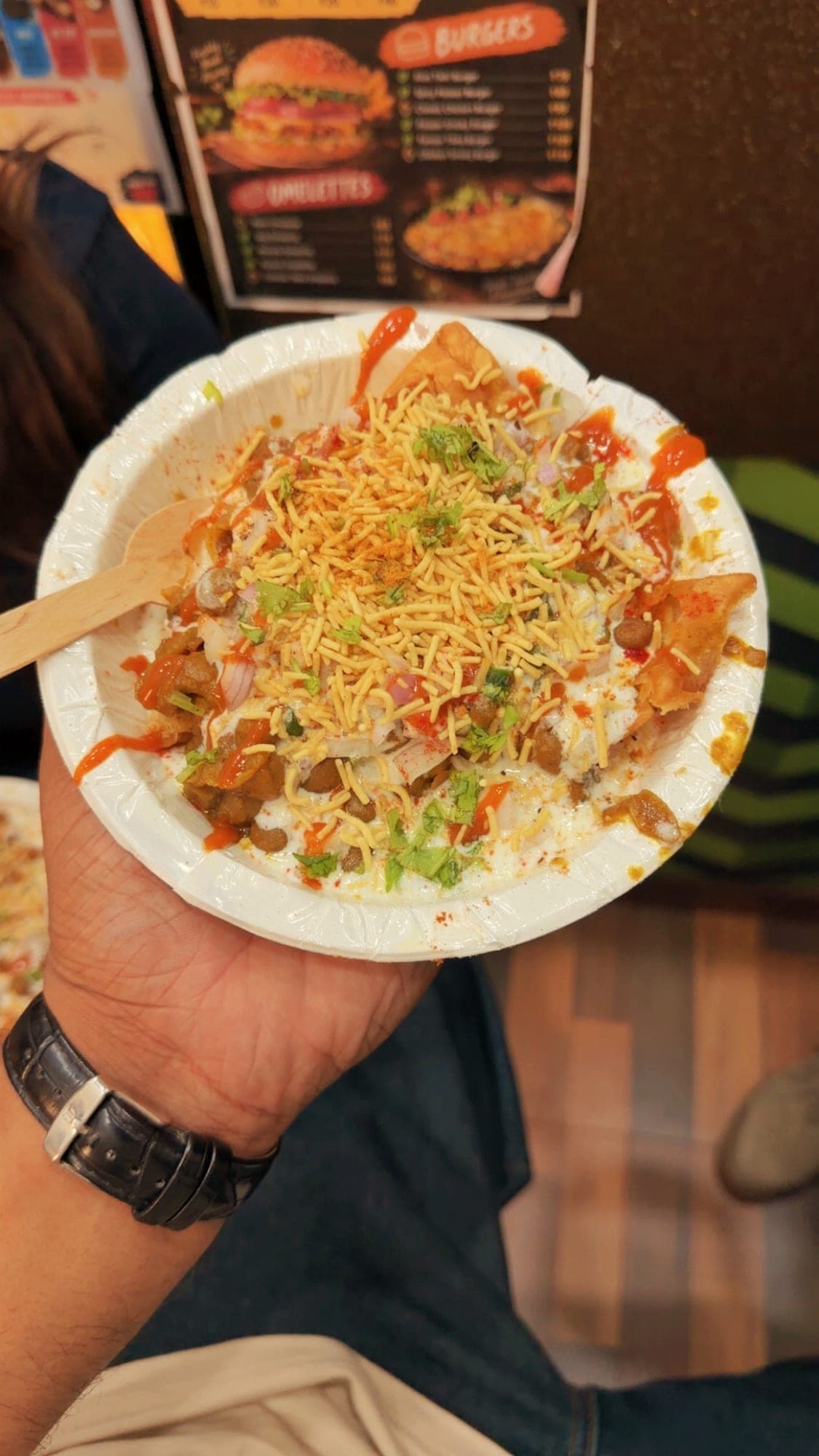 A hand holding a white paper bowl of samosa chaat with a wooden spoon: pieces of samosa and chickpeas under curd, red sauce, chopped onion, coriander and a heap of fine sev, with a menu board showing burgers and omelettes behind