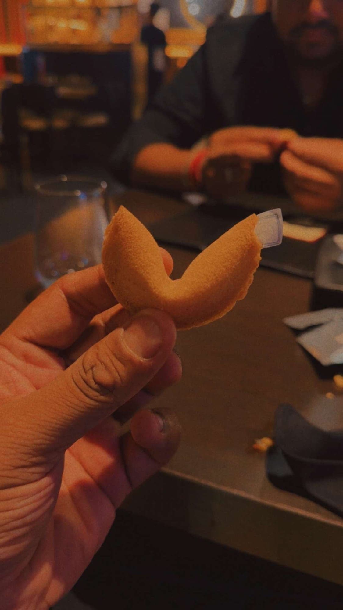 A hand holding a cracked fortune cookie with a small paper slip showing, in a dim restaurant with a blurred table and diner behind