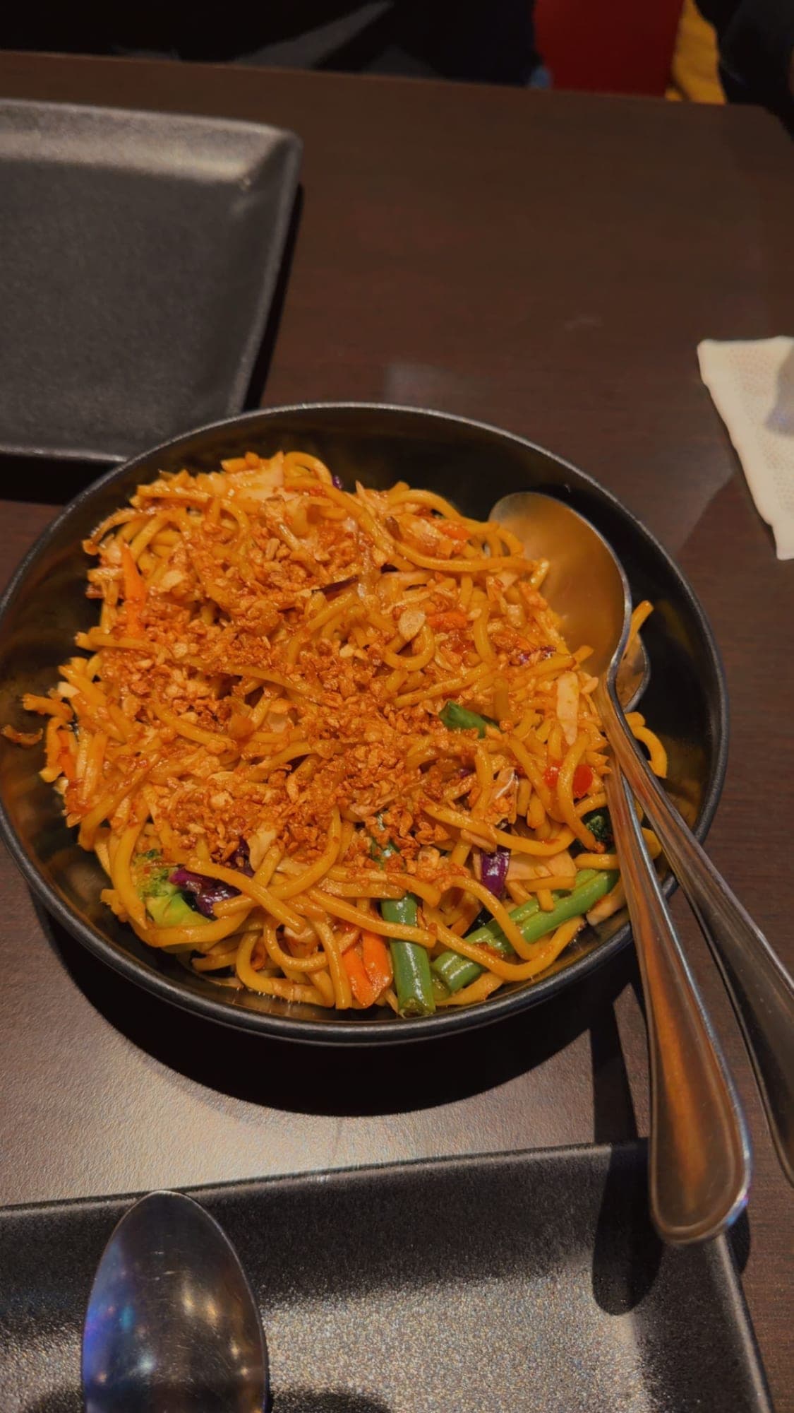 A black bowl of stir-fried noodles with green beans, carrot and cabbage, covered in golden crispy garlic, with a serving spoon and fork resting in it