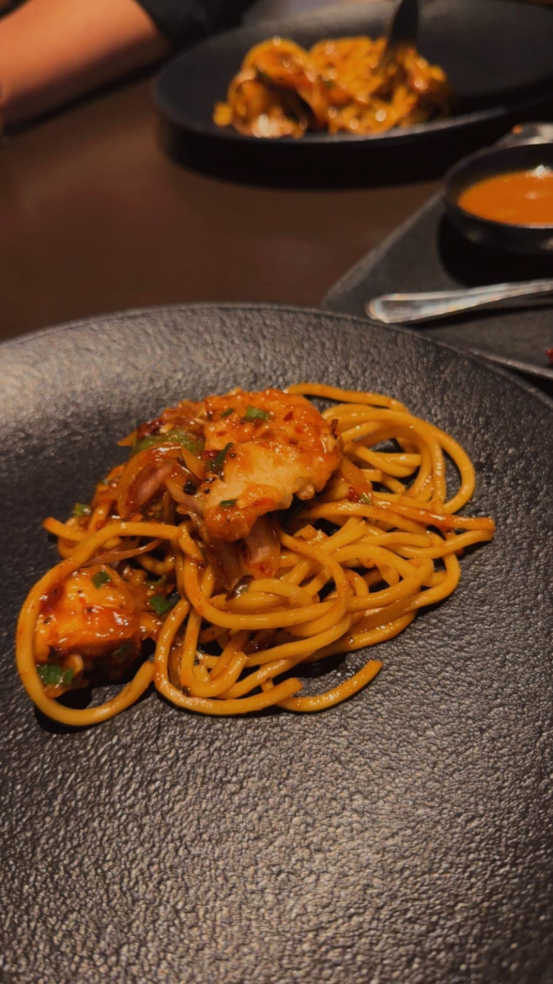 A swirl of golden noodles with sticky glazed pieces and spring onion on a dark speckled stone plate, with another plate blurred behind
