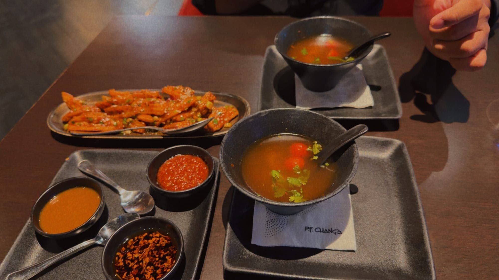 A dark table from above: two black bowls of orange soup with coriander and cherry tomatoes on a P.F. Chang's napkin, a tray of three chilli sauces, and a long dish of crisp glazed batons with peas