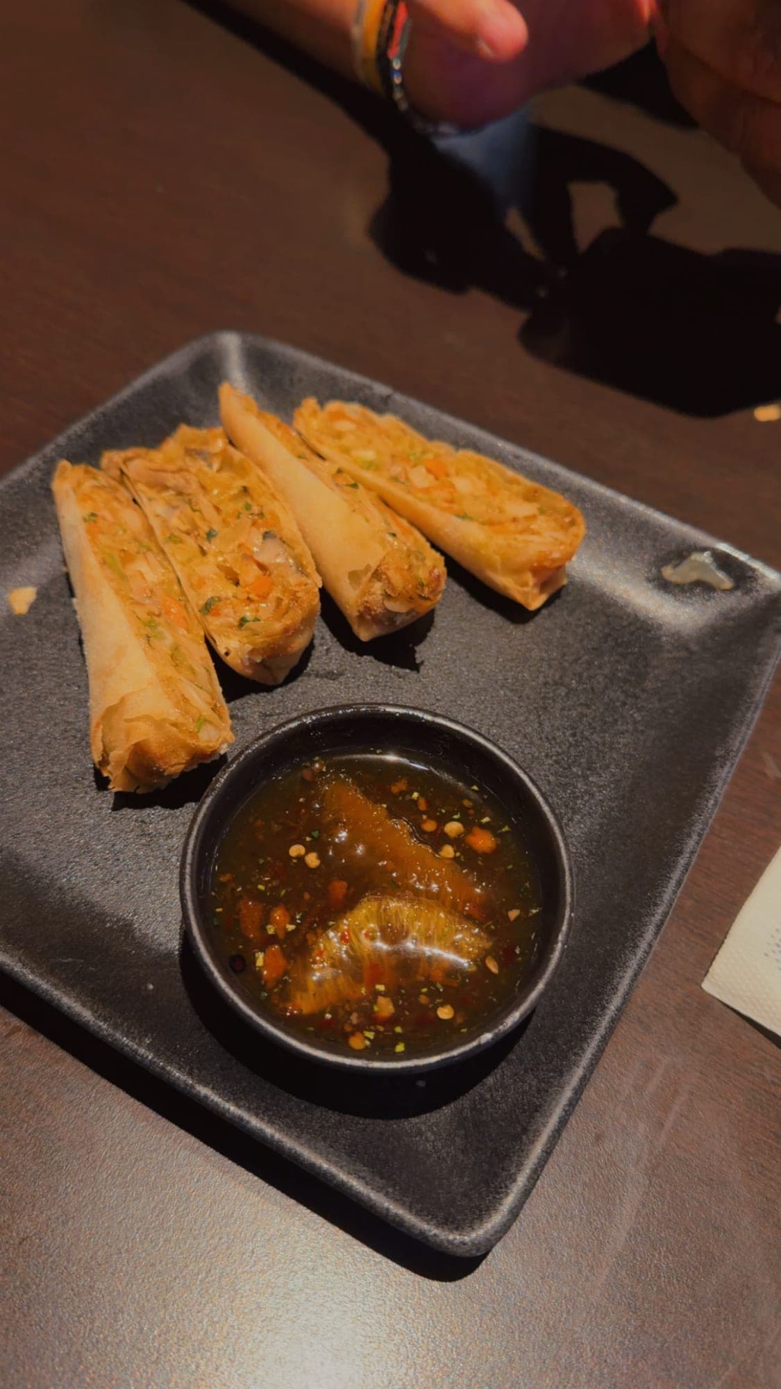 Four crisp spring rolls cut in half on a square black plate, with a small black bowl of dark chilli dipping sauce beside them