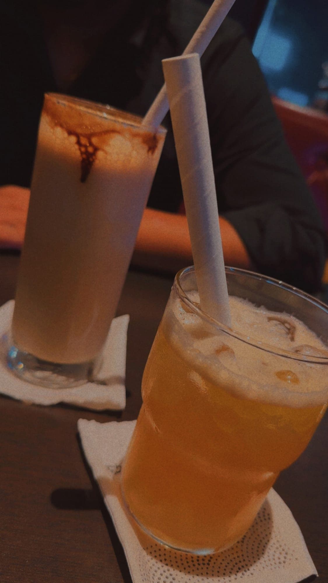 Two tall glasses with paper straws on white napkins: a creamy drink streaked with chocolate and an amber iced drink, in warm dim light