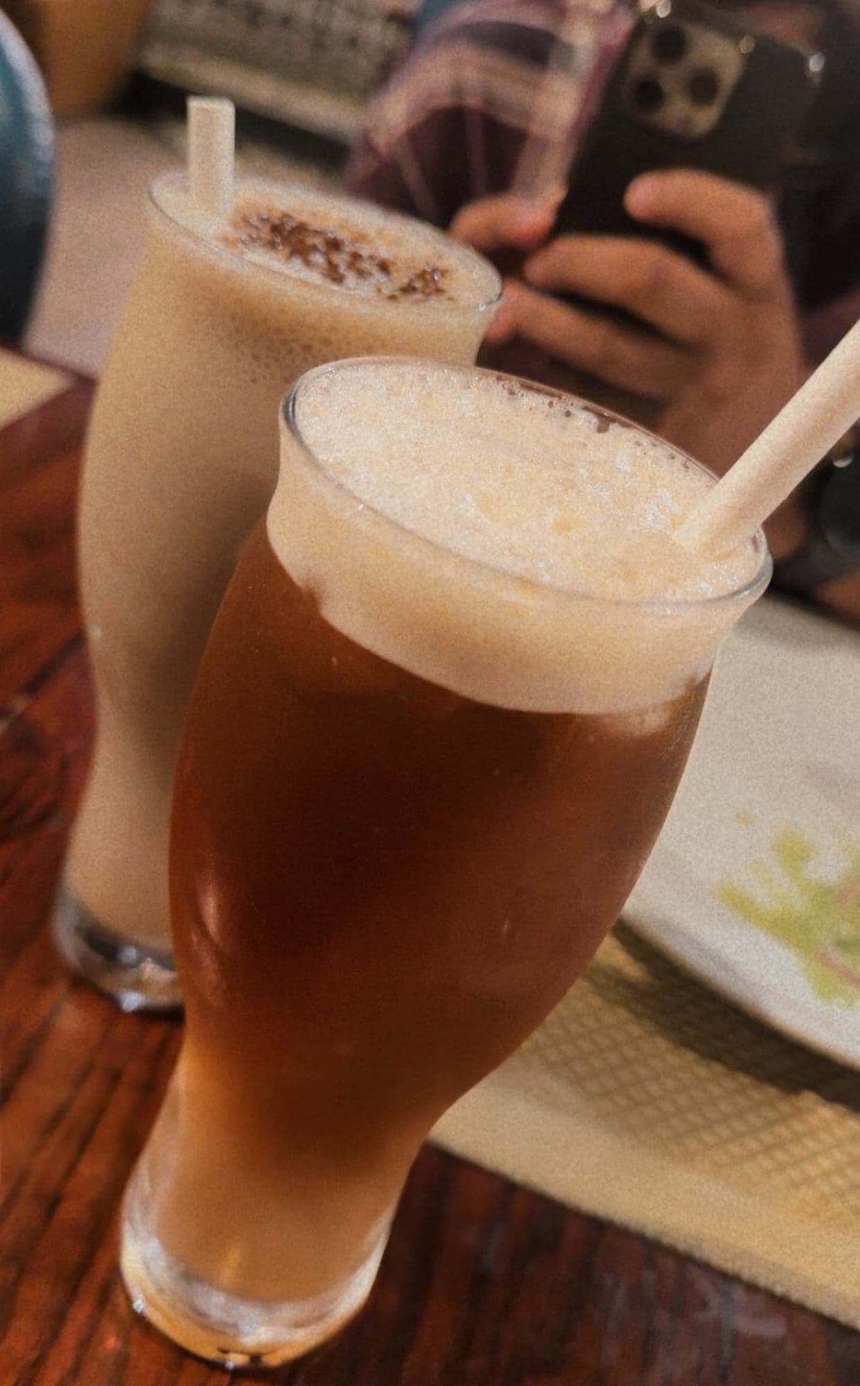 Two tall glasses on a wooden table: a peach iced tea with a thick pale foam and a paper straw in front, and a cold coffee dusted with brown powder behind it, with a hand holding a phone out of focus above