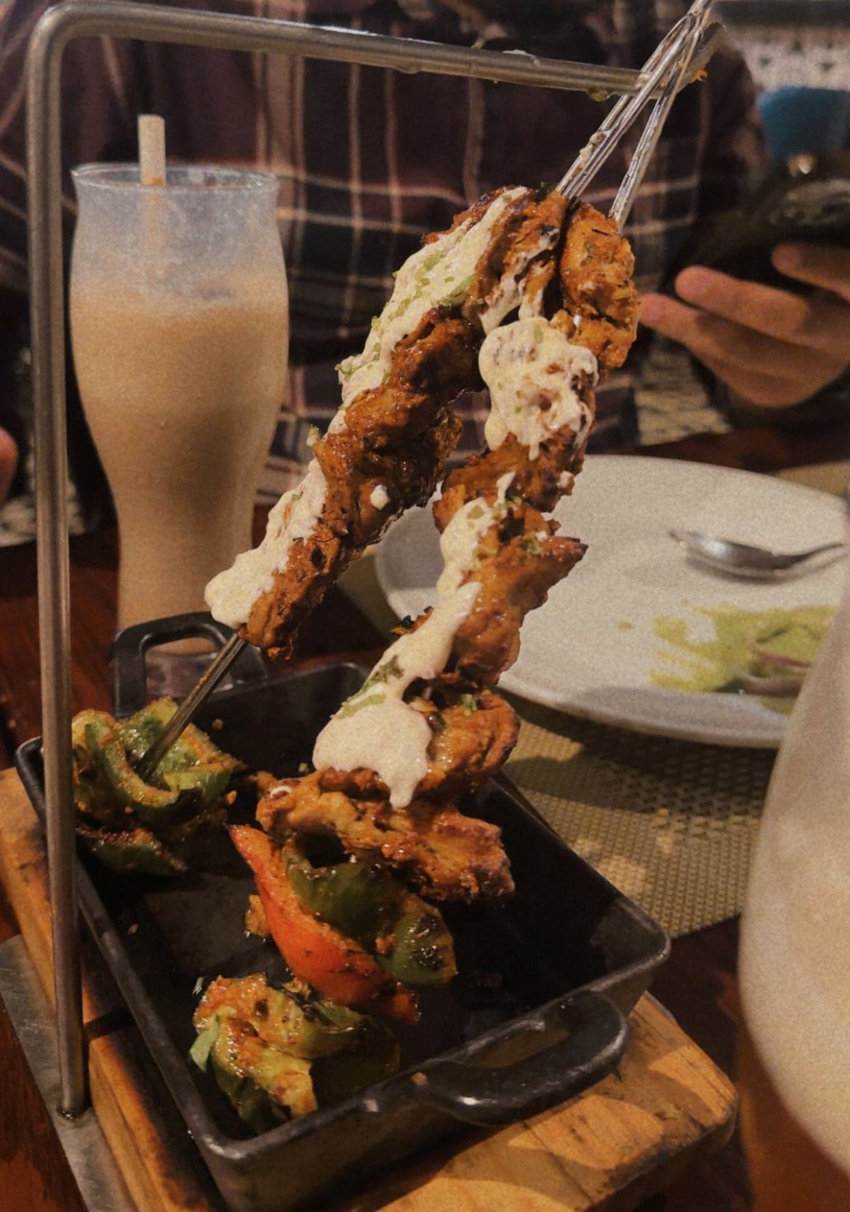 Pieces of grilled malai chaap on two skewers hanging from a metal stand over a cast-iron tray of charred peppers, with a drizzle of white cream sauce, and a tall cold coffee behind