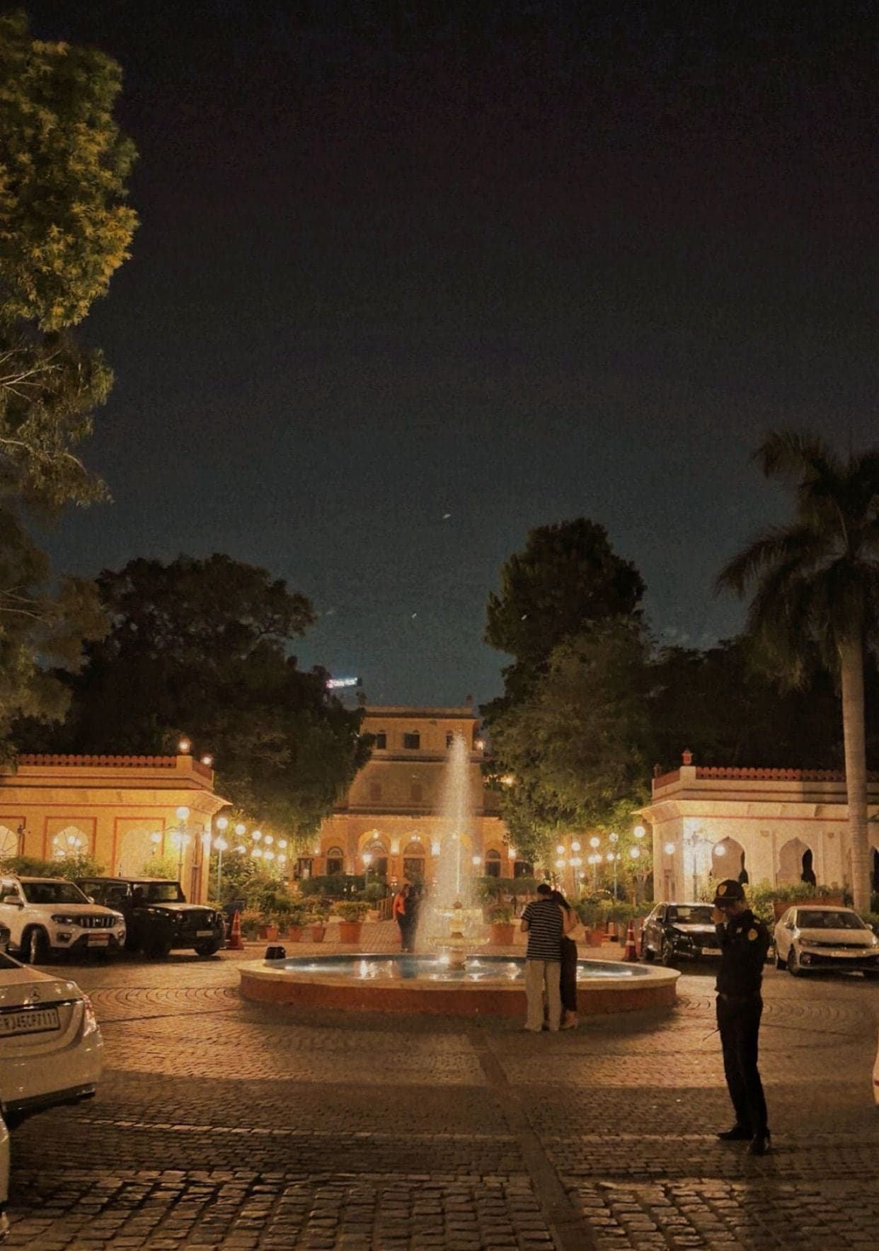 A cobbled courtyard at night with a lit fountain in a round pool, a warmly lit palace building with arched windows behind it, low pavilions with strings of lamps on either side, parked cars, a palm tree and two people standing by the fountain