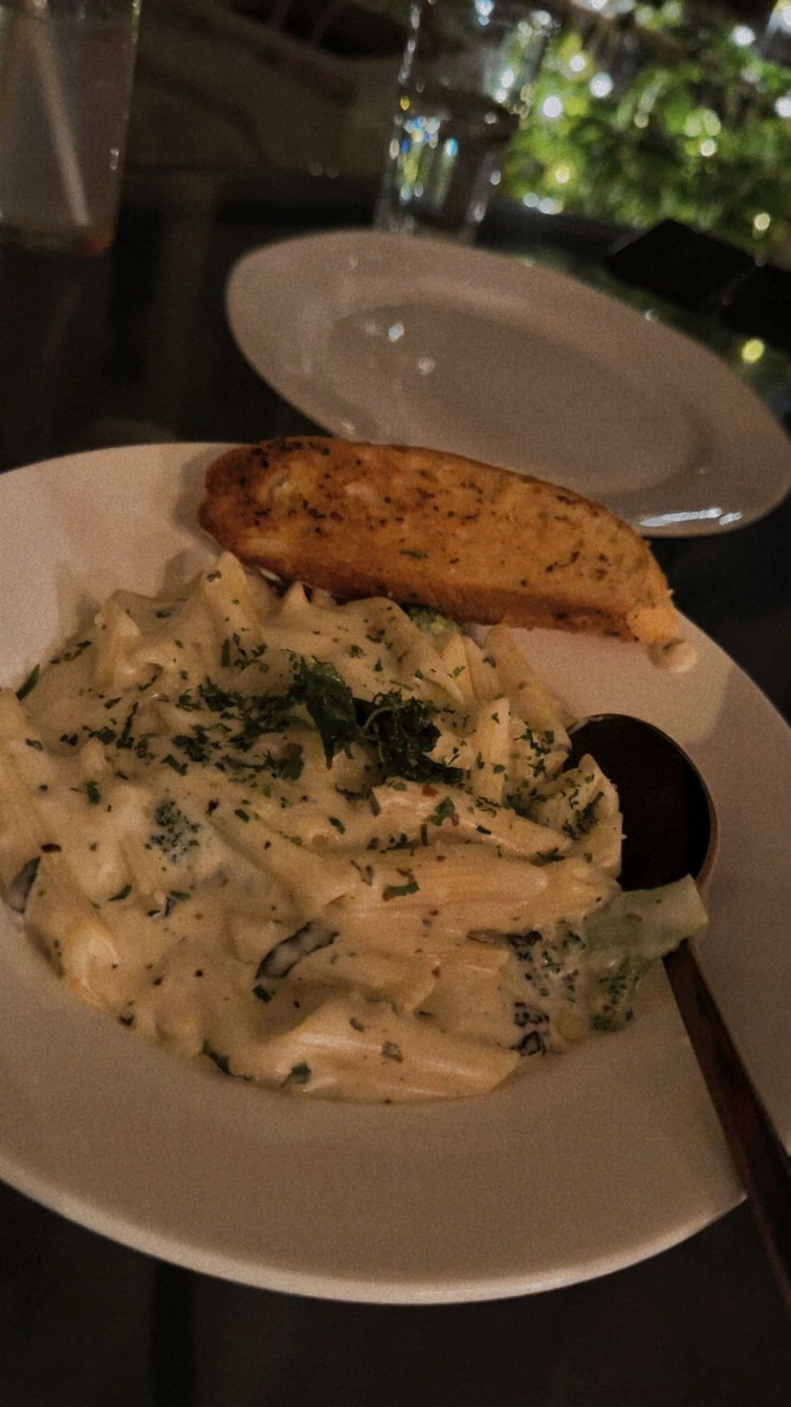 A wide white bowl of creamy penne with broccoli and chopped herbs, with a slice of toasted garlic bread on the rim and a dark spoon, on a dimly lit table with an empty plate behind