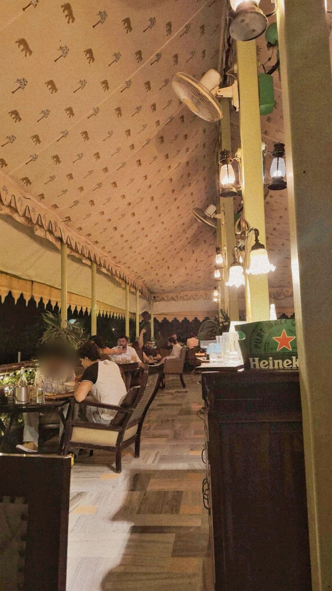 A tented dining area at night with a ceiling printed with gold elephants and palm trees, wall fans and lanterns on pale green posts, people eating at wooden tables, and a green ice bucket on a dark wooden cabinet in the foreground
