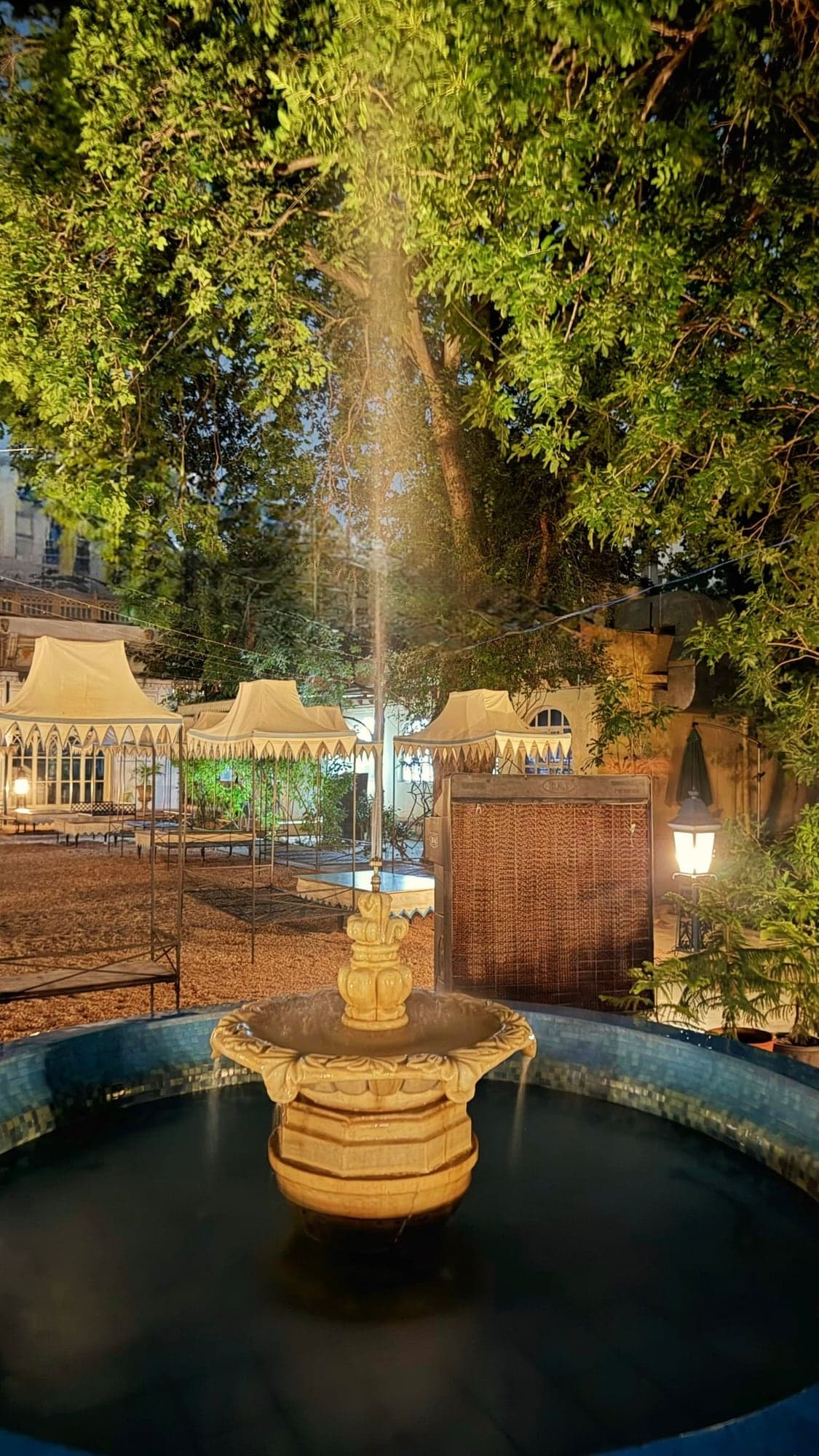 A tiered cream stone fountain in a round pool edged with blue mosaic tiles, a thin jet of water rising through the leaves of a large tree, with cream tent canopies, a gravel yard and a glowing lamp behind, all looking brightly lit although the scene was almost completely dark