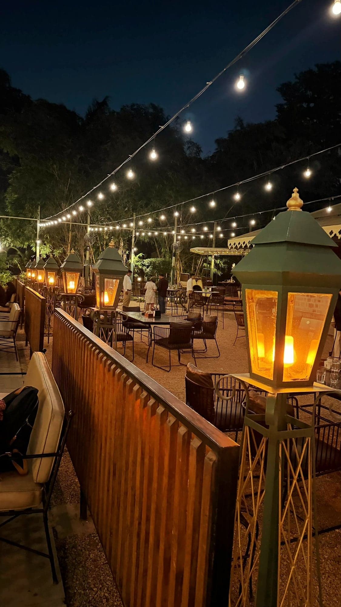 A gravel garden dining area at night with strings of round bulbs overhead, green lanterns glowing with a warm flame light along a wooden slatted fence, black metal chairs and tables, a few people standing in the distance and tall trees behind