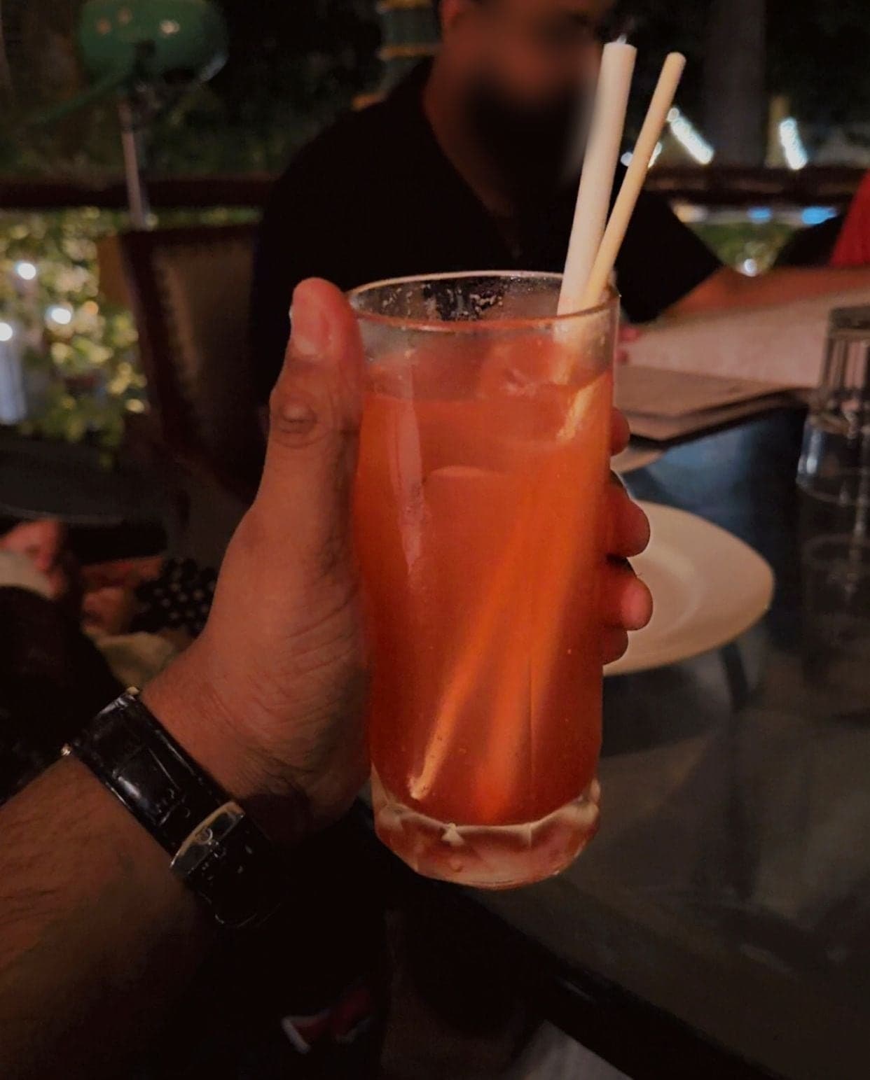 A hand with a black-strapped watch holding a tall glass of orange-pink mocktail with two paper straws, on a dimly lit restaurant table with a plate and glasses behind