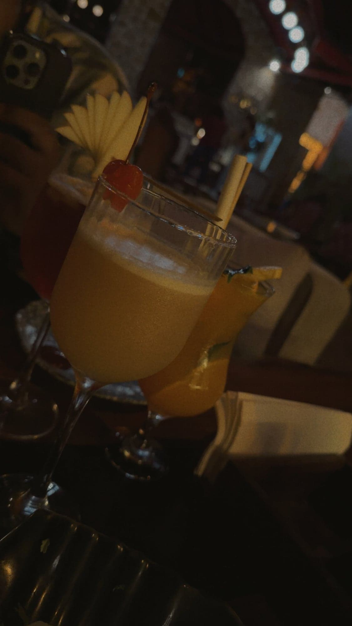 A stemmed glass of pale orange drink with a cherry on a pick and a fan of sliced apple, next to a bright orange drink with a lime garnish and a red drink behind, in a dim bar with warm lights