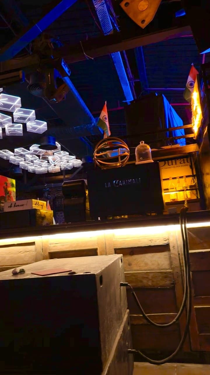 The ceiling and bar of a dark venue: beams and ducts lit blue, clusters of glowing glass cubes hanging at the left, two small Indian flags above a black espresso machine, and shelves of bottles lit amber at the right, over a wooden panel with a warm strip of light