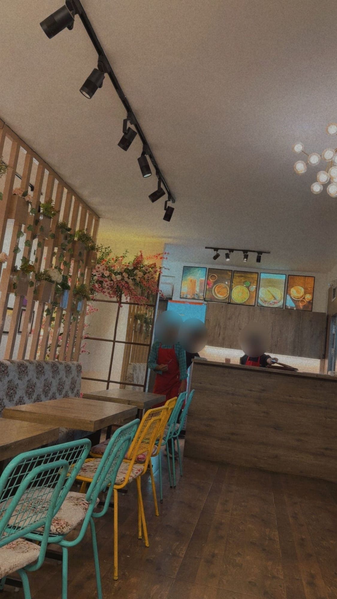 Inside a small café: teal and yellow wire chairs with floral cushions at wooden tables, a slatted wooden divider with plants and pink flowers, and a counter at the back under lit food pictures, with staff whose faces are blurred