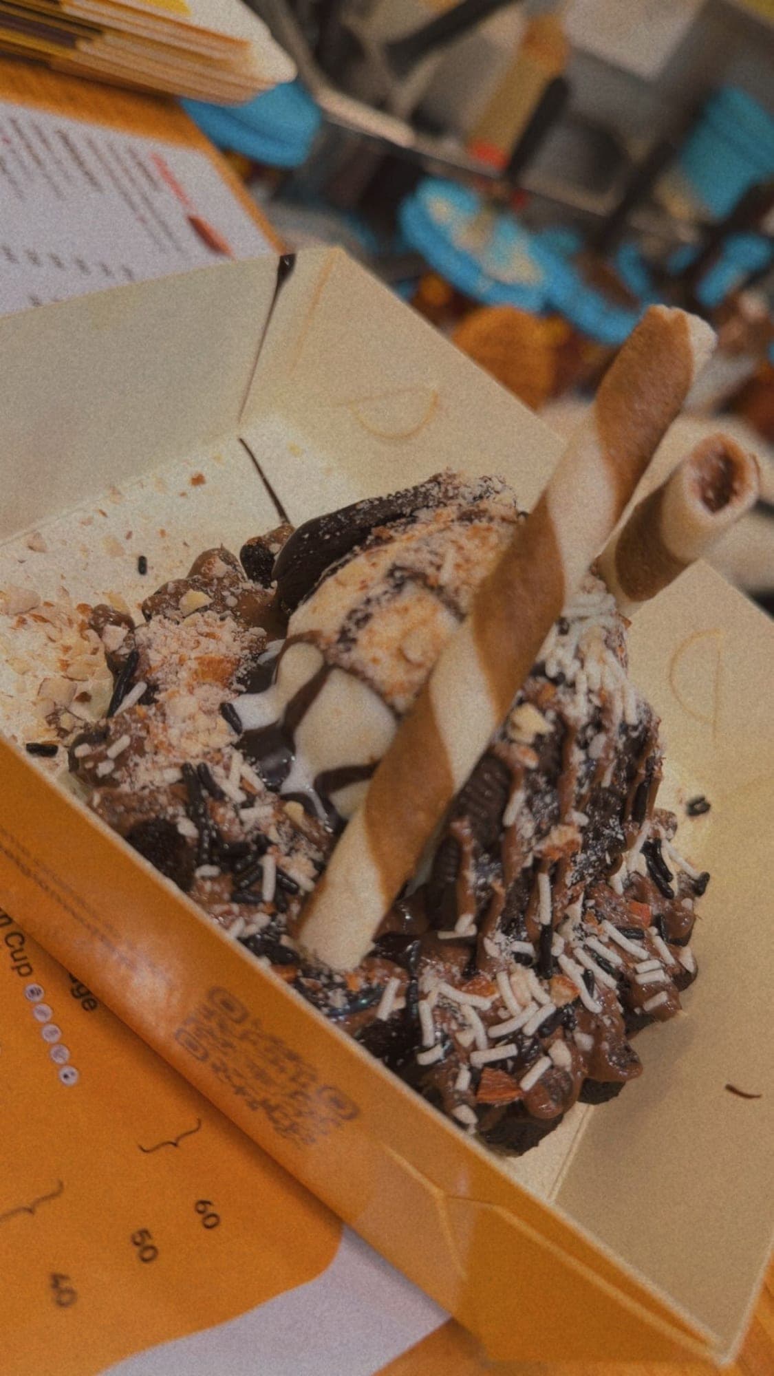 A yellow takeaway box holding a waffle buried under chocolate sauce, crushed nuts, white and dark sprinkles and ice cream, with two striped wafer rolls standing on top