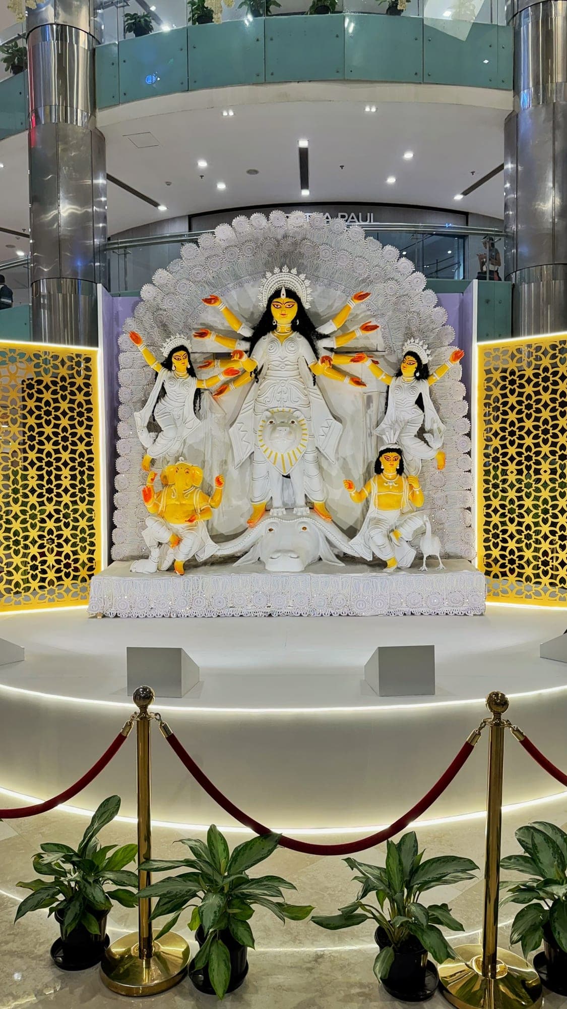 A large white idol of the goddess Durga with many raised arms, standing on a lion above a buffalo, with four smaller figures around her including Ganesha, in front of a white lace-like arch and between two golden patterned screens, on a round stage ringed with light behind red rope barriers and potted plants, in a mall atrium