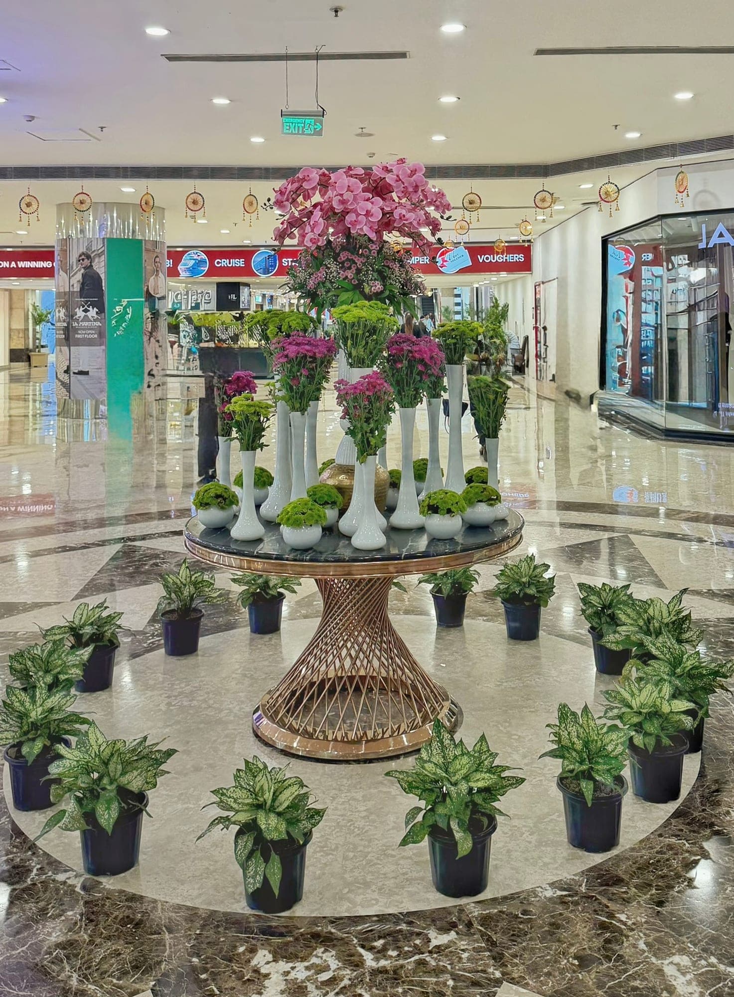 A round table with a twisting rose-gold base holding tall white vases of pink orchids, green and magenta chrysanthemums, ringed by black pots of variegated green plants on a polished marble floor, with shop fronts and hanging festive decorations in the mall behind