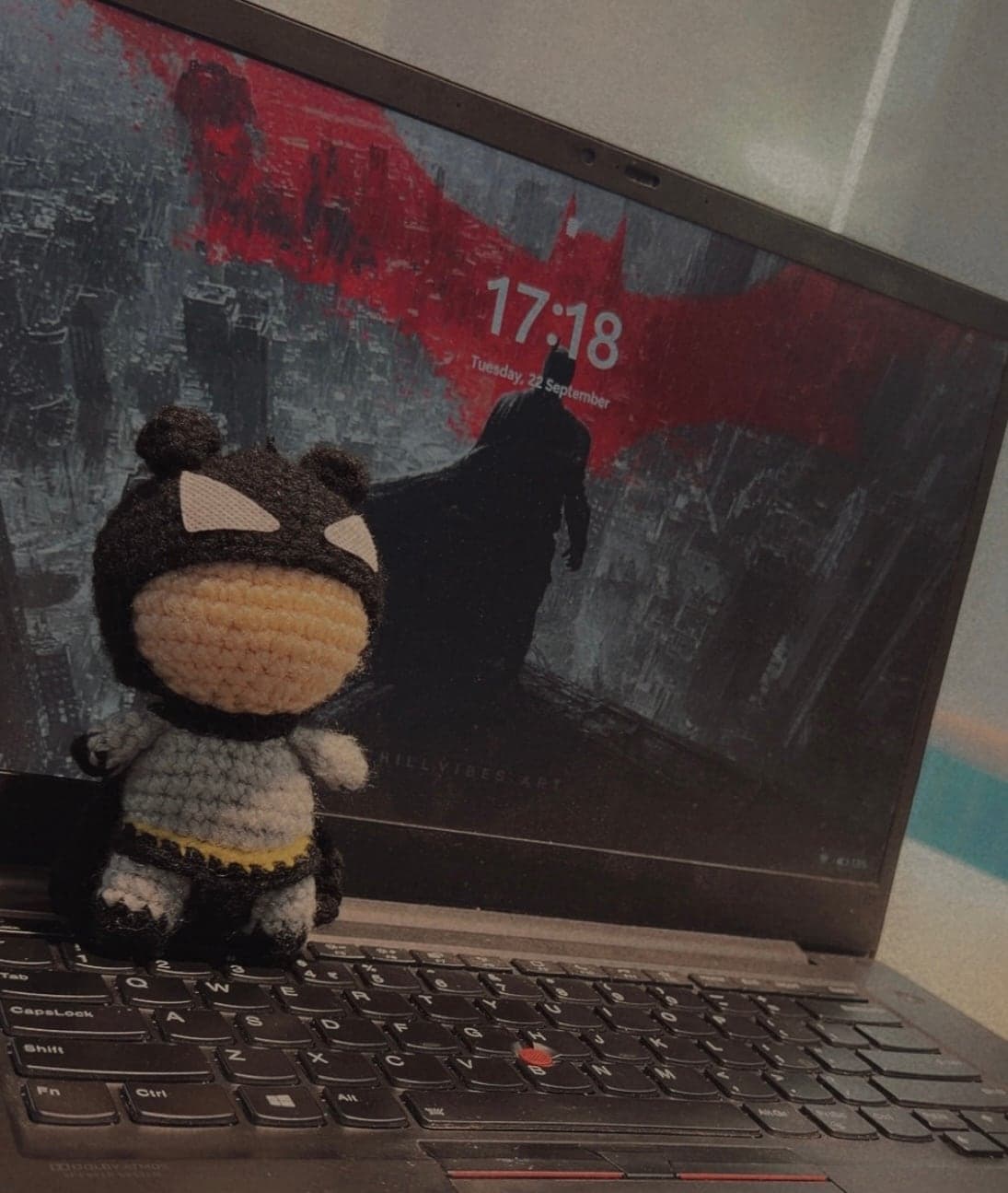 A small crocheted Batman doll in a dark cowl with white eyes, a pale yellow face, a grey body and a yellow belt, standing on the keyboard of an open laptop whose screen shows a Batman lock-screen wallpaper with a red bat symbol and the time 17:18