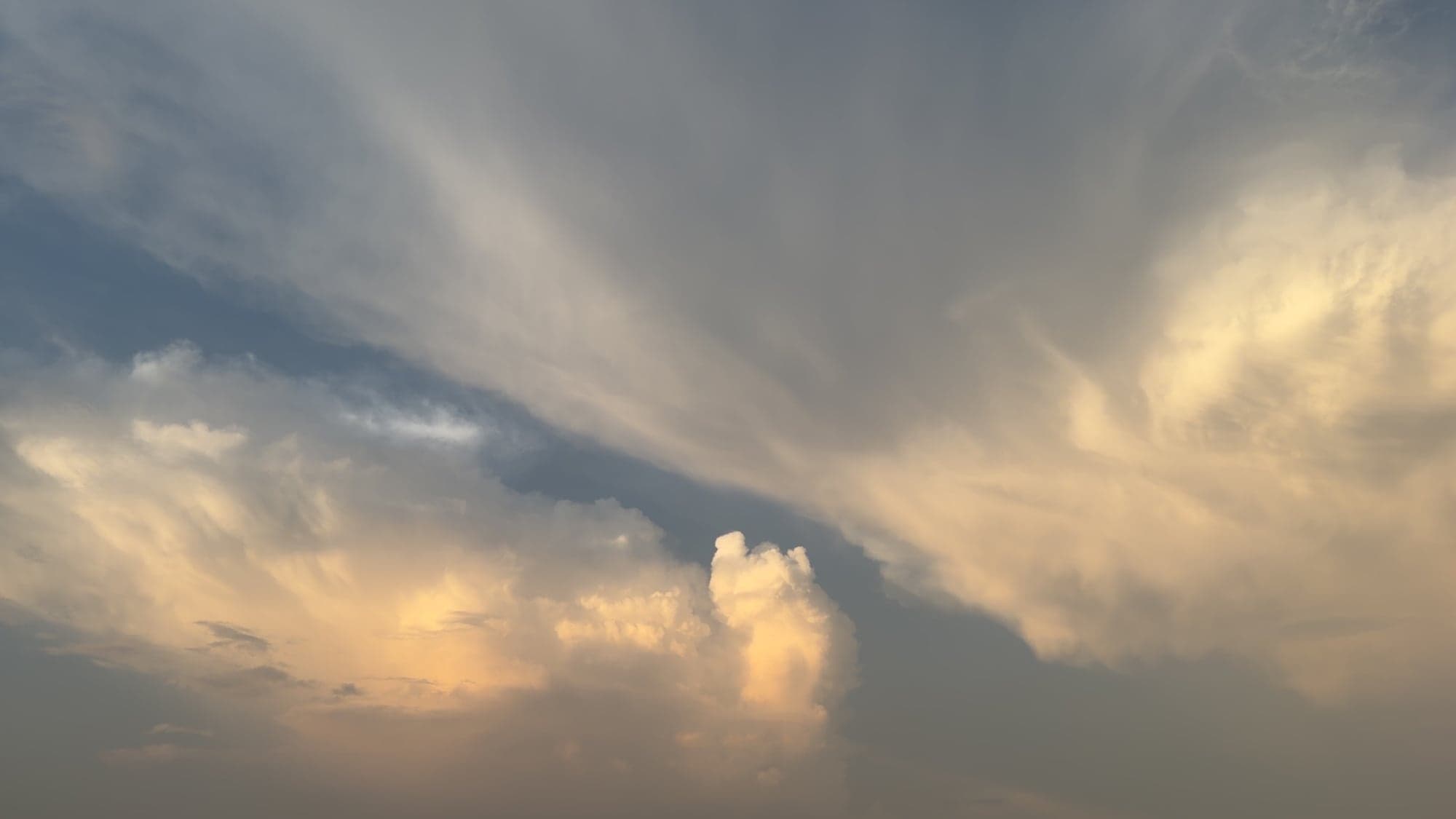 A sunset sky with a golden-lit cumulus tower rising from a bank of peach clouds at the bottom, under long streaks of grey and blue cloud