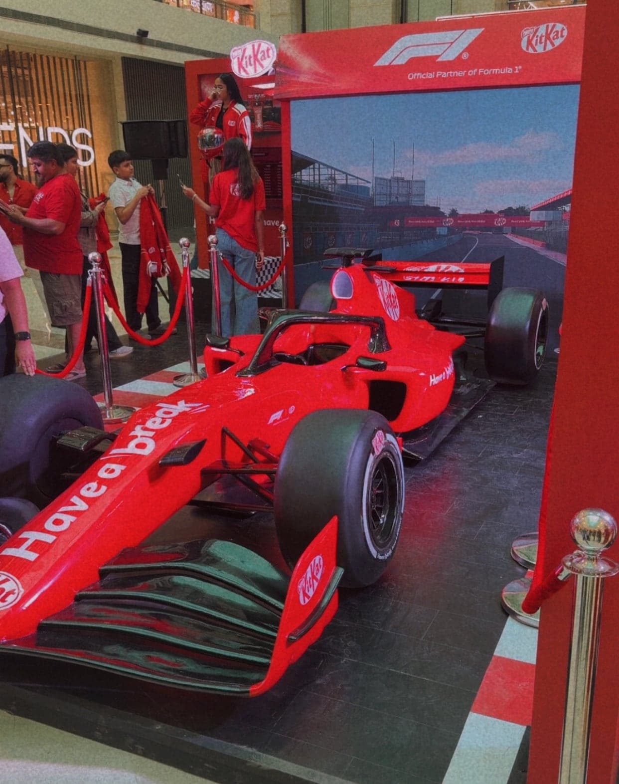 A bright red Formula 1 style car on a display stage, with the KitKat logo and the words Have a break along its nose and sides, in front of a backdrop of a race track and a red banner reading Official Partner of Formula 1, with rope barriers and staff in red shirts at the left