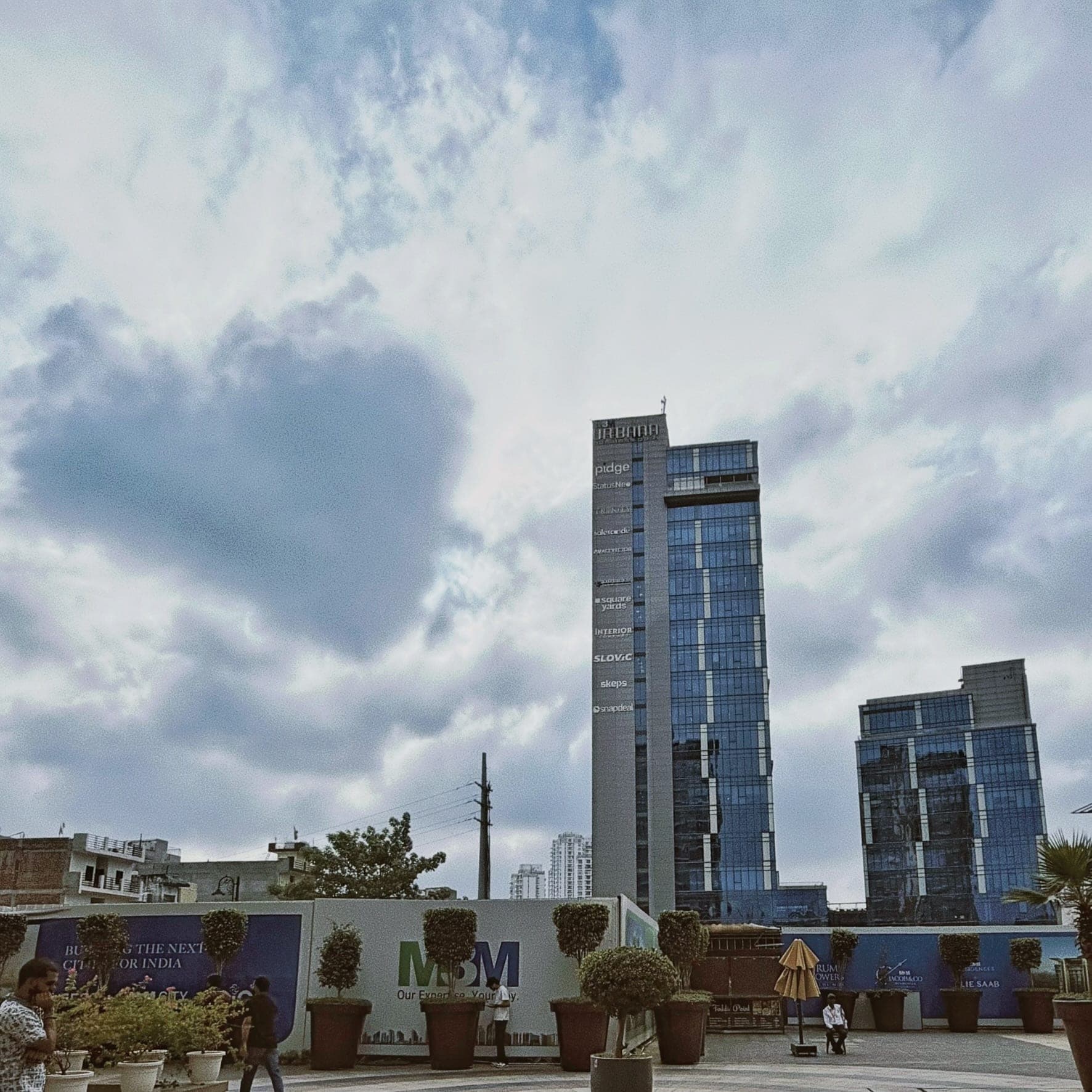 A heart-shaped patch of dark blue cloud among white and grey clouds, above the glass towers of an office park with an Urbana sign, over a plaza with potted trees, hoardings and a few people walking