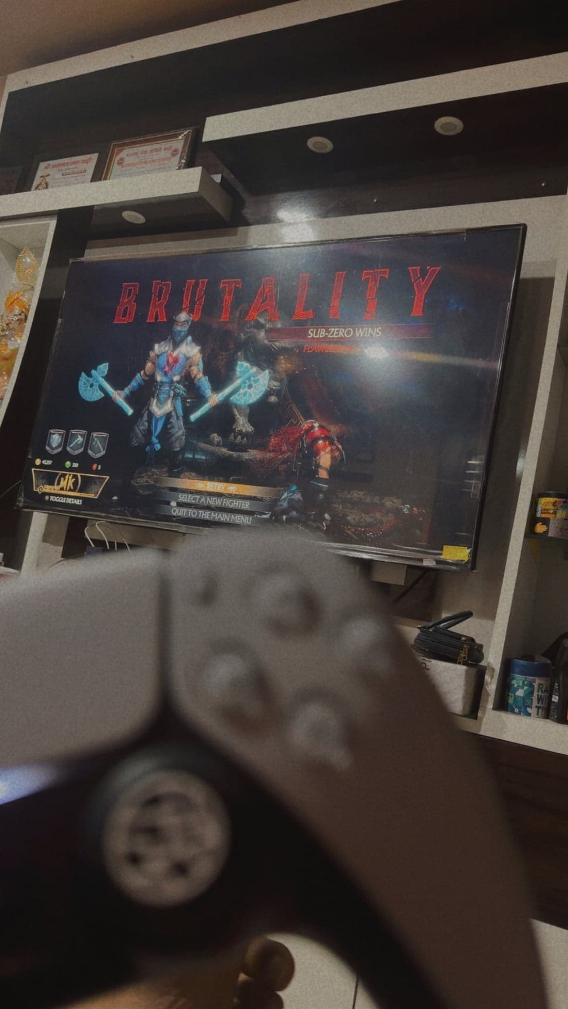A blurred PS5 controller in the foreground and a wall-mounted TV behind it showing the Mortal Kombat Brutality screen, with Sub-Zero holding two ice axes and the words Sub-Zero wins
