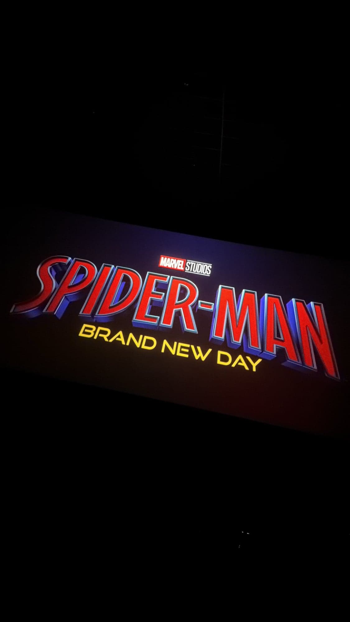 The Marvel Studios title card for Spider-Man: Brand New Day glowing in red, blue and yellow on a dark cinema screen, tilted in the frame