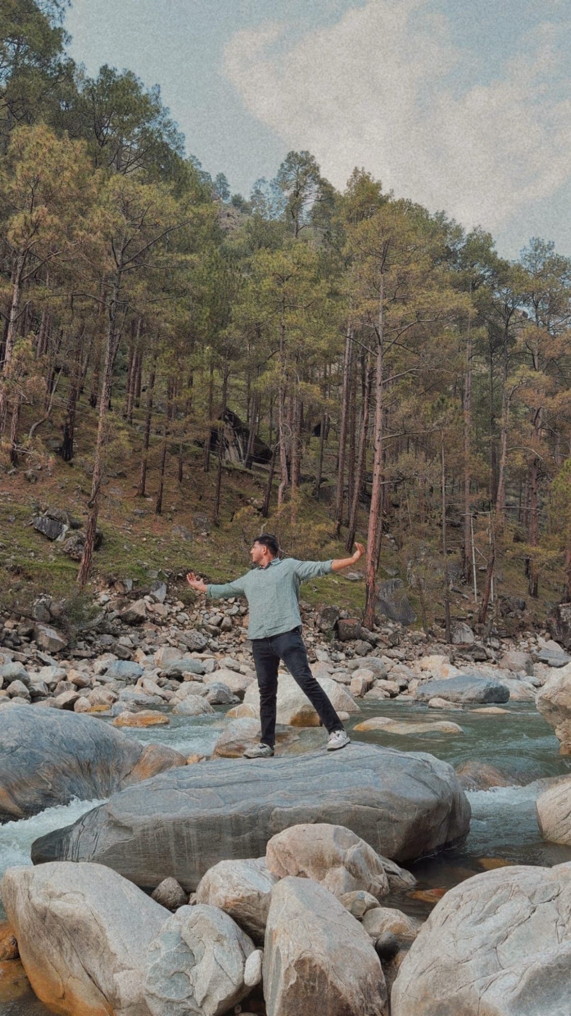 A person in a pale green shirt standing on a large grey boulder in a river, arms stretched out to the sides, with a pine-covered hillside behind and a soft, grainy, faded colour grade
