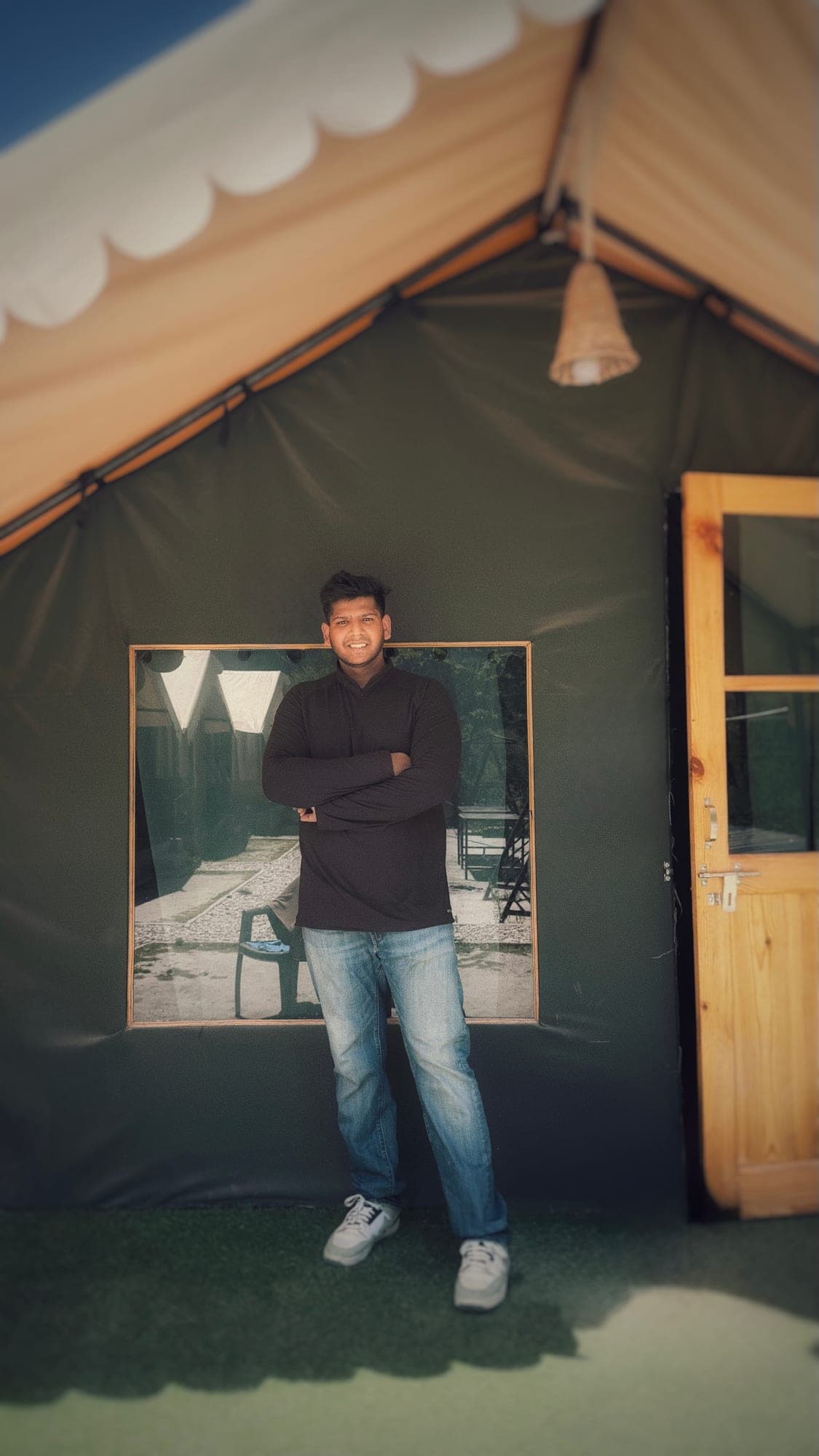 A smiling person with folded arms, in a dark top and jeans, standing against the green canvas wall of a tent, beside a pine door and under a scalloped tan canopy with a woven lamp hanging from the ridge