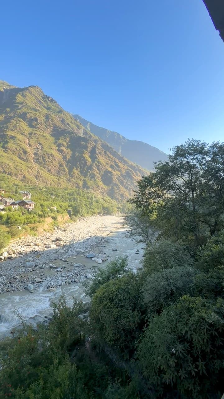 A bend of the Parvati river in a green valley seen from above, its stony bed and white water running between trees on the right, with a few houses on the far slope and steep sunlit mountains under a clear blue sky