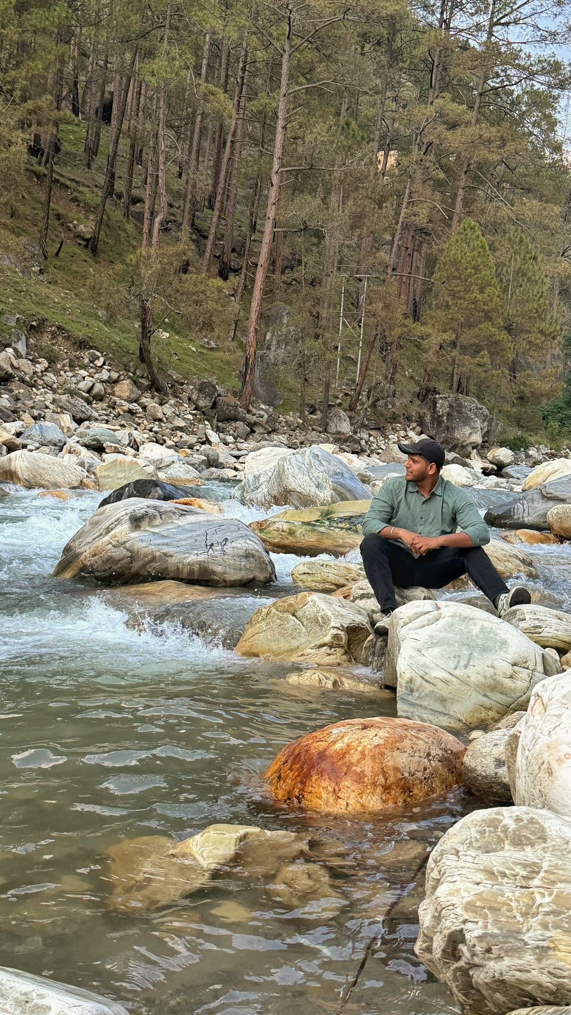 A person in a green shirt and black cap sitting on a pale boulder in the middle of a fast mountain river, looking away to the side, with a pine-covered slope behind