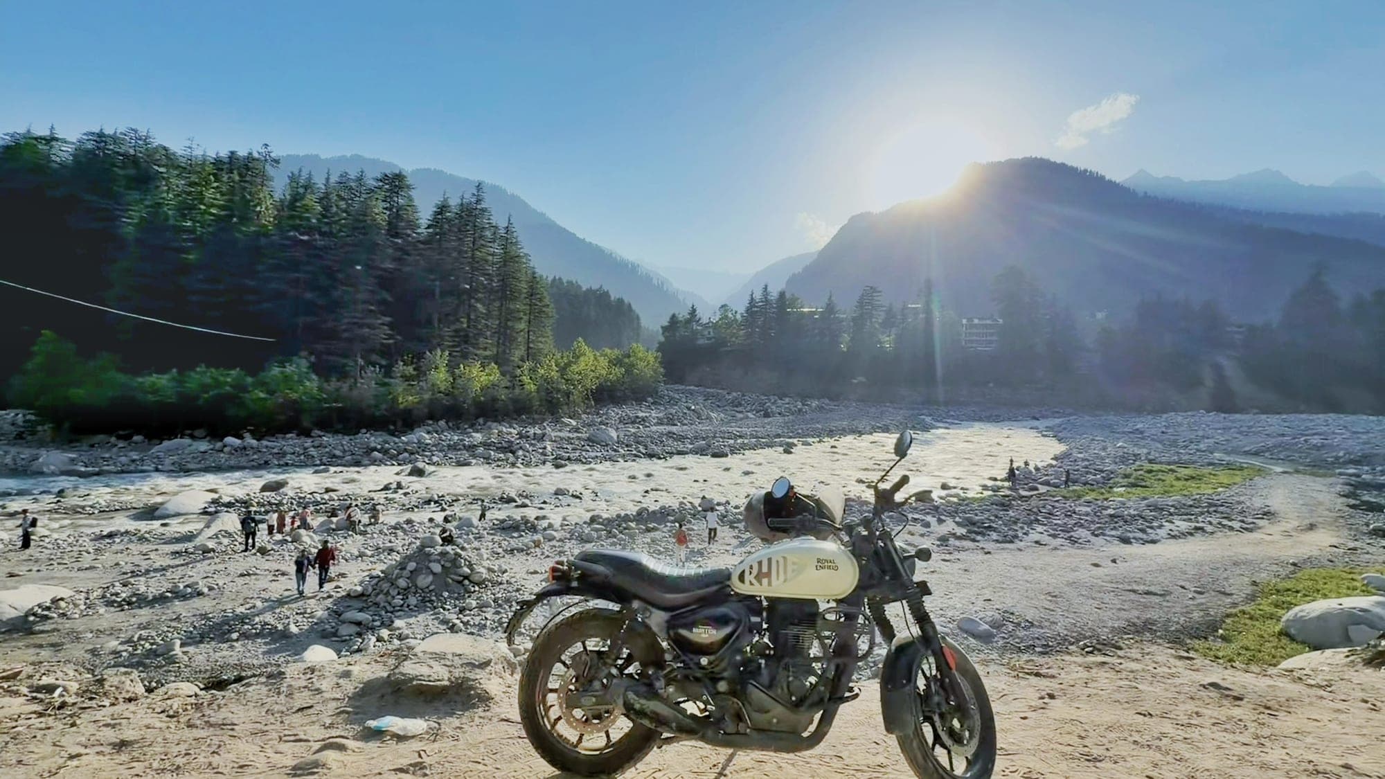 A cream and black Royal Enfield motorcycle parked on a sandy riverbank in the foreground, with a wide braided river, a few people walking on the stones, a dark pine forest and the sun flaring over a mountain ridge in the background