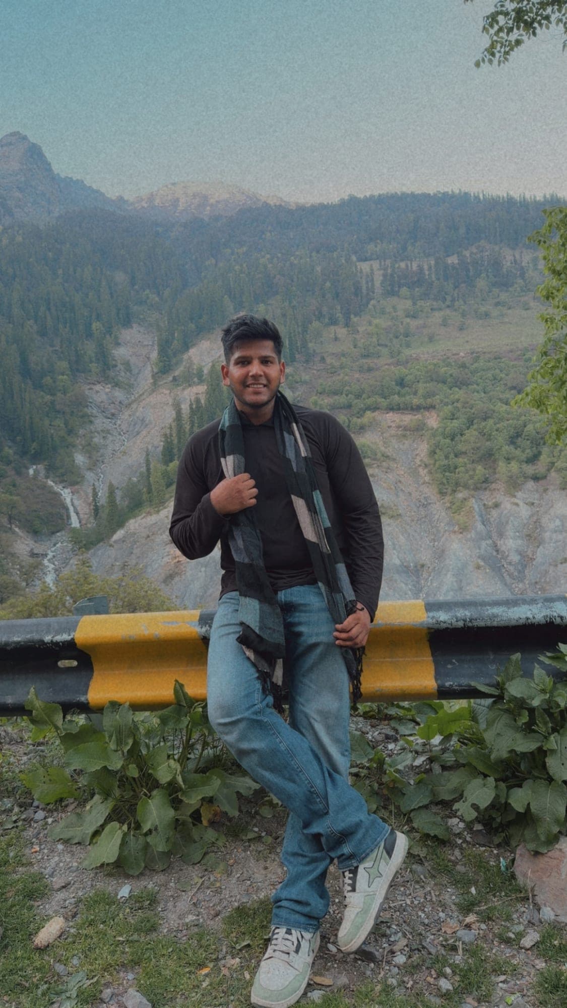 A smiling person in a black top, jeans and a checked scarf leaning on a yellow and black crash barrier at the roadside, with a forested mountain slope and a thin waterfall behind in a hazy, grainy blue-green grade